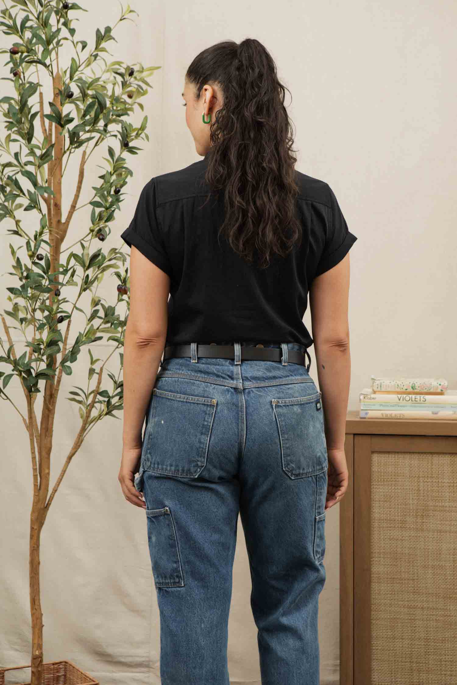 A person with long, curly hair in a ponytail stands facing away, wearing the Bridge & Burn Bea Shirt in Black and blue jeans. A tall plant is on the left and a small table with books sits on the right.