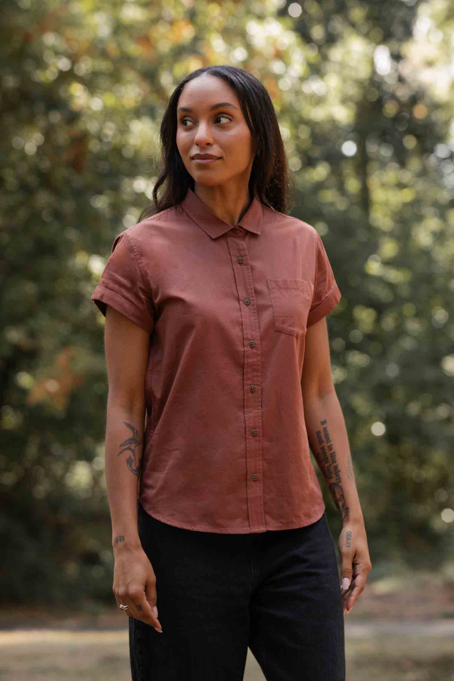 A person with long dark hair wears the Bridge & Burn Bea Shirt in Copper and black pants, standing outdoors looking to the side, with softly blurred green trees and sunlight in the background.