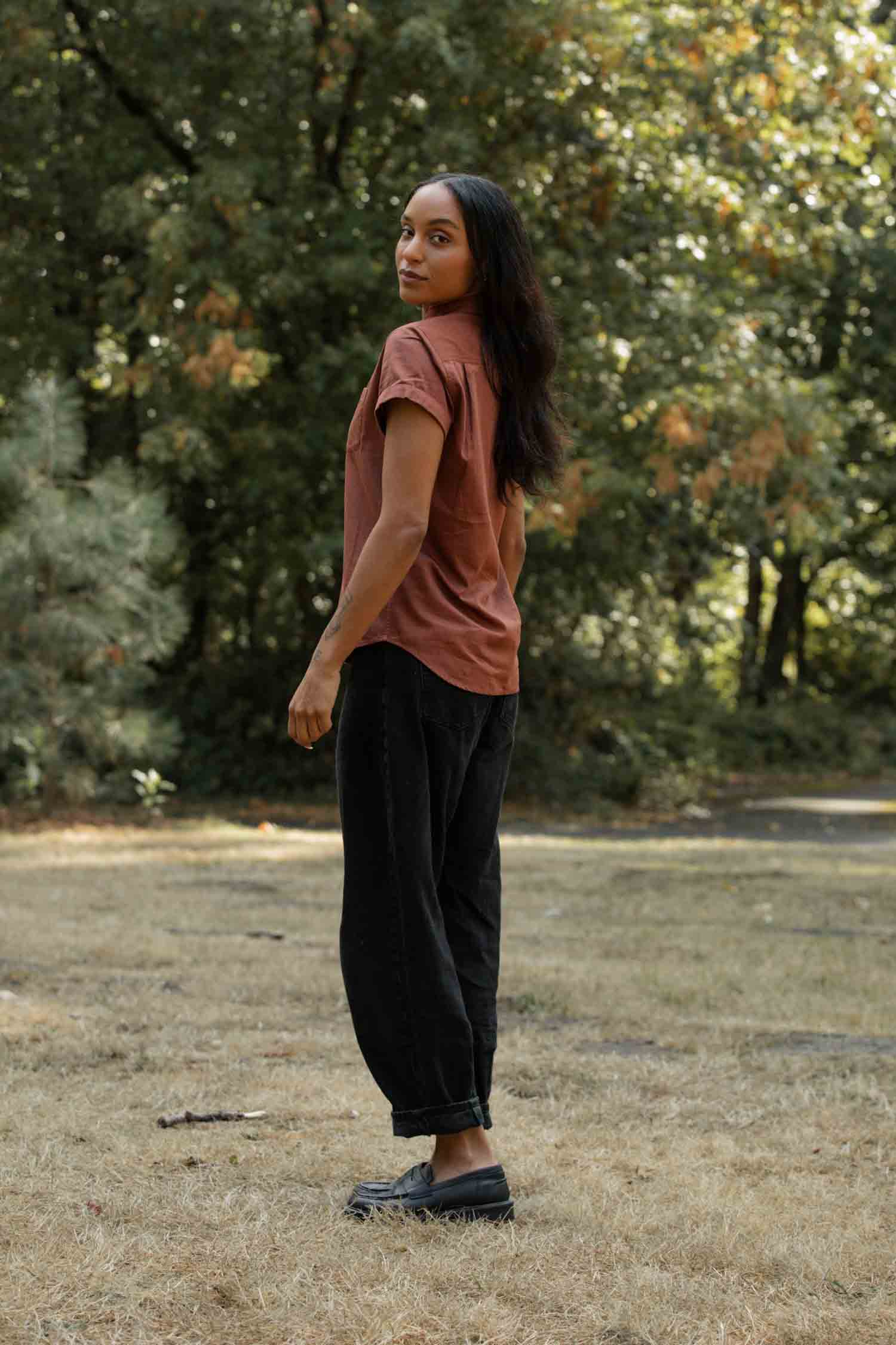 A person with long dark hair stands outdoors on dry grass, looking over their shoulder at the camera. They wear the Bridge & Burn Bea Shirt in Copper (organic cotton, tailored fit), black pants and black shoes, with trees behind.