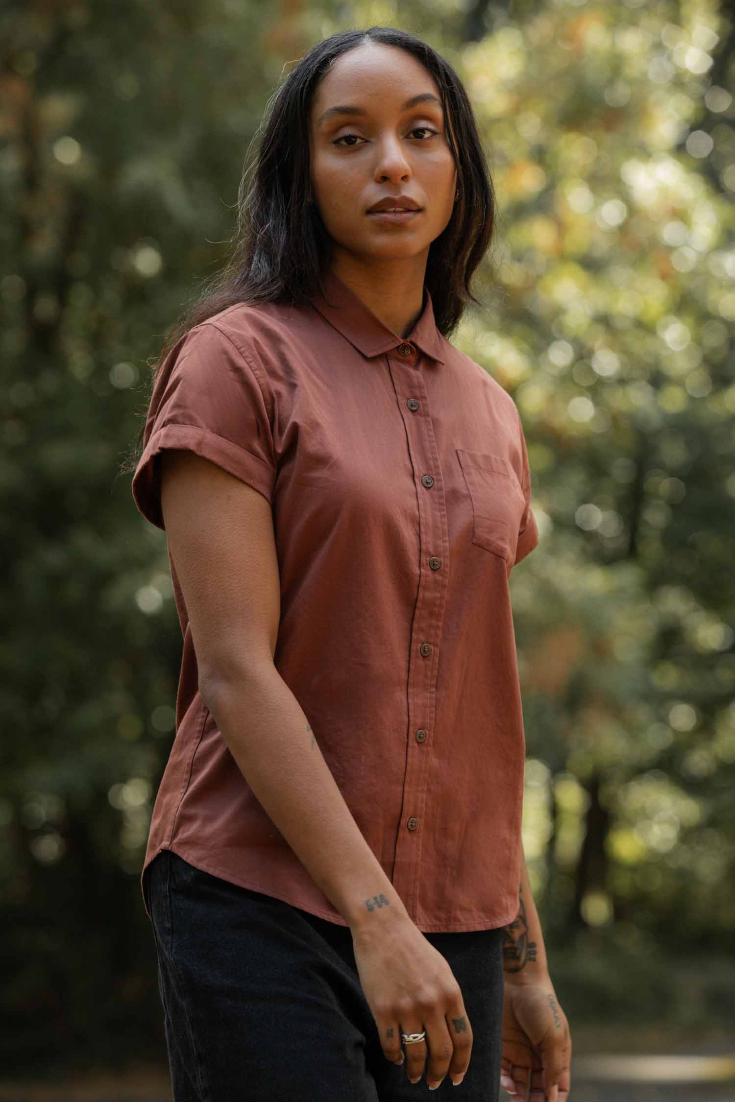 A woman with long dark hair wears the Bridge & Burn Bea Shirt in Copper—an organic cotton short-sleeve button-down—standing calmly in a sunlit green wooded area, looking at the camera.
