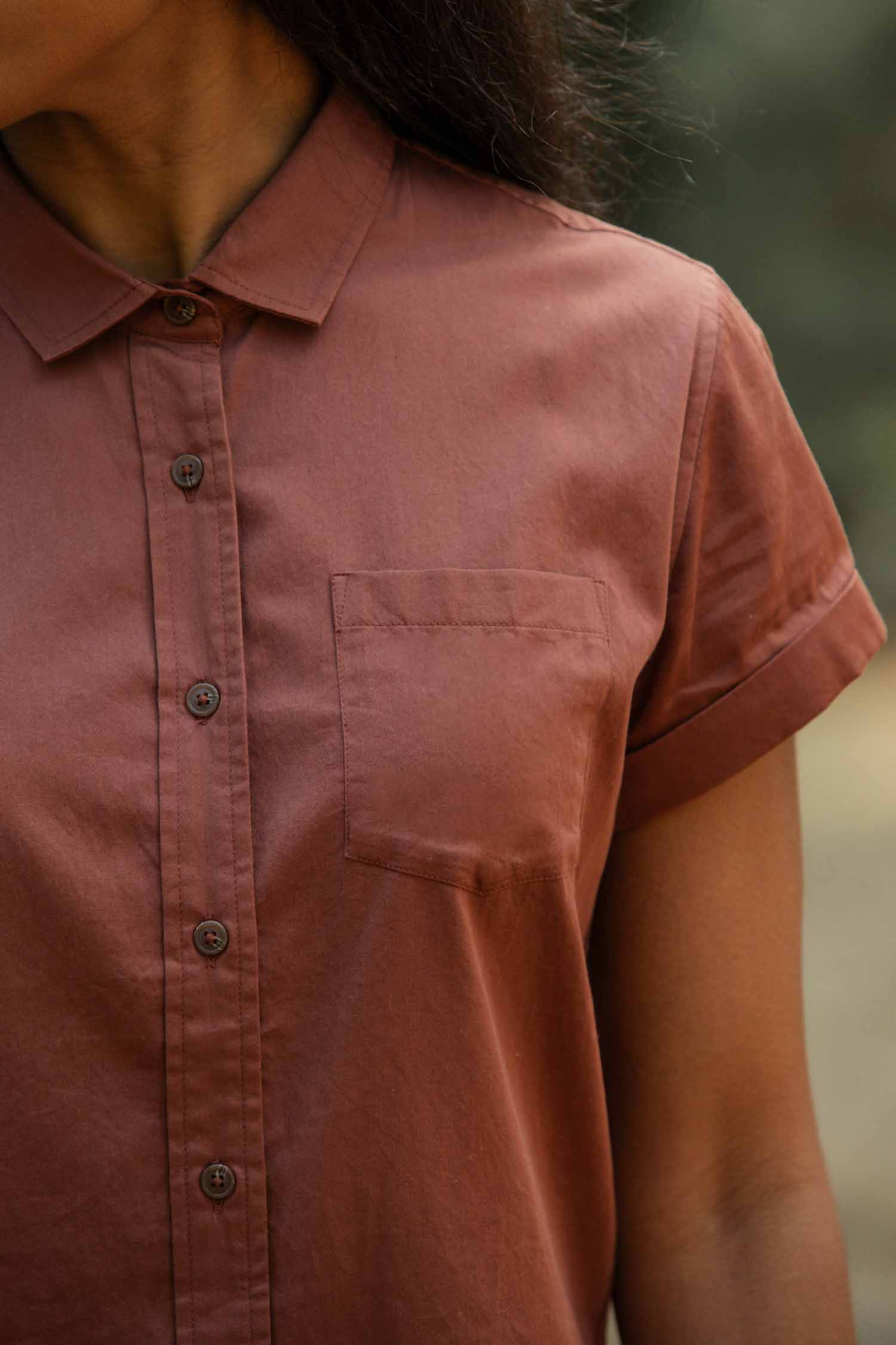 A person wears the Bridge & Burn Bea Shirt in Copper, a tailored fit short-sleeve button-down with a chest pocket. Only the upper torso and part of the face are visible, set against a blurred background.
