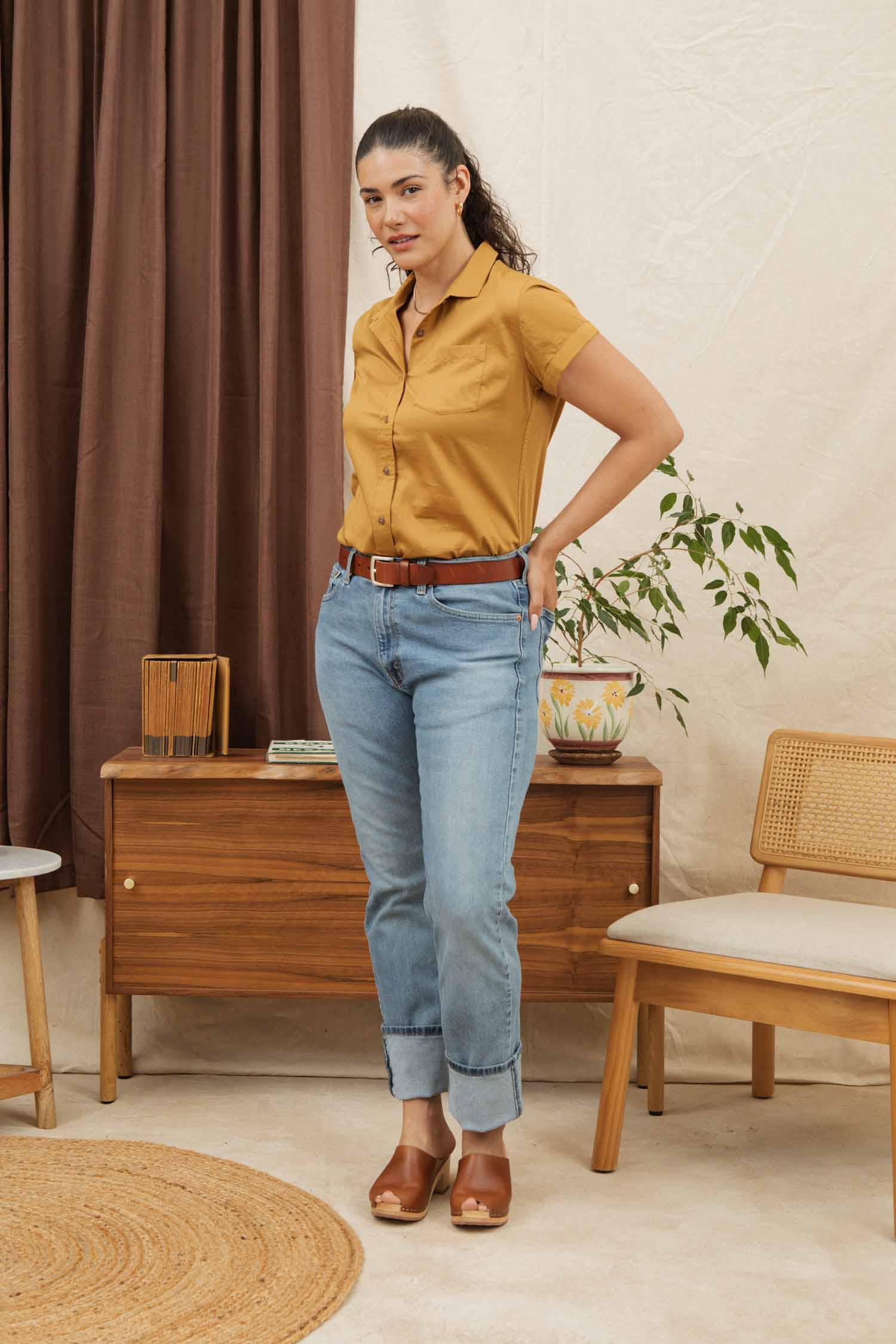 A woman wears the Bridge & Burn Bea Shirt in Gold, paired with light blue jeans and brown heels, standing in a modern room featuring wooden furniture, a plant, and neutral-colored curtains.