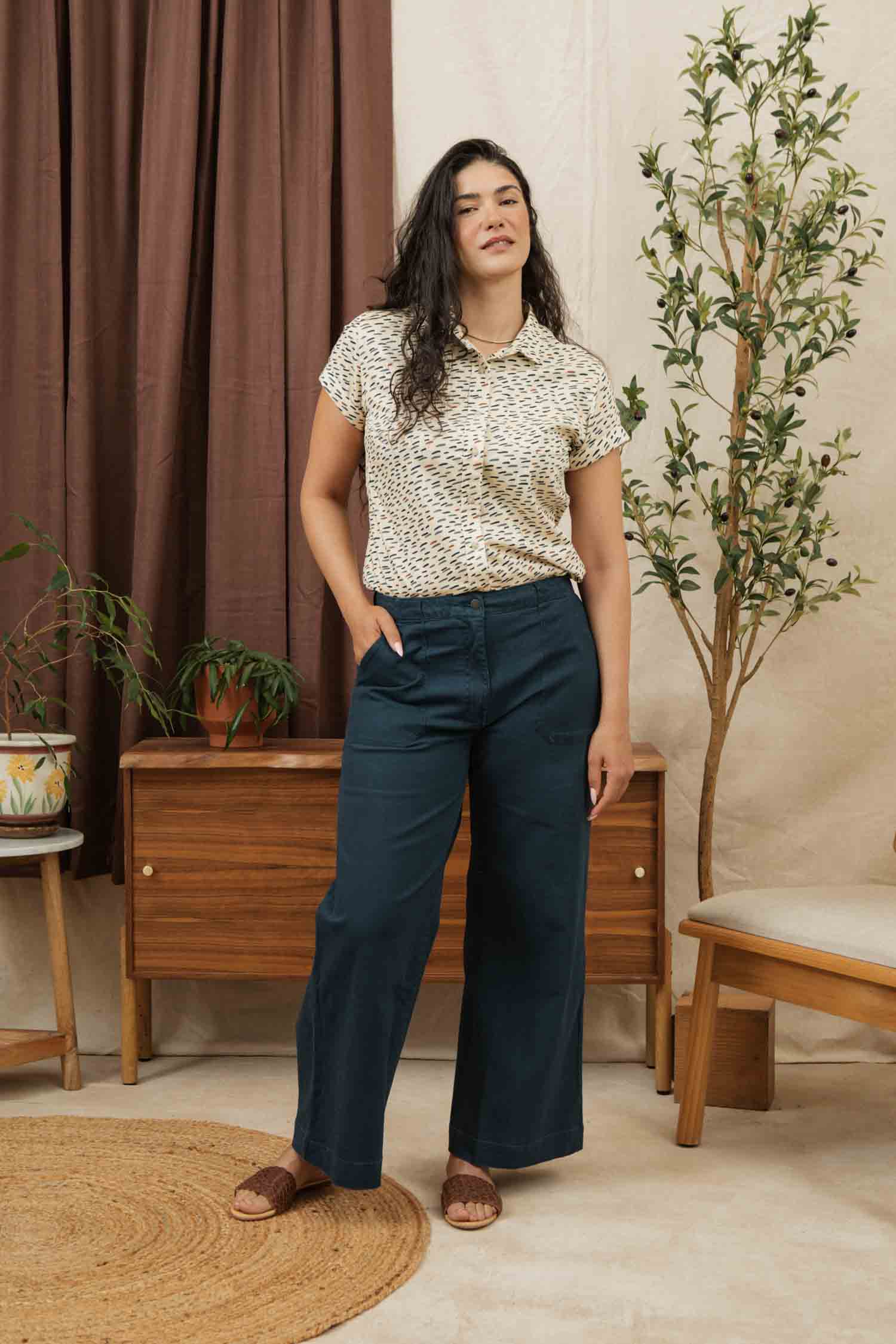 A woman with long dark hair stands indoors, wearing the Bridge & Burn Bea Shirt in Ivory Dashes. She is surrounded by wooden furniture, potted plants, and brown curtains.