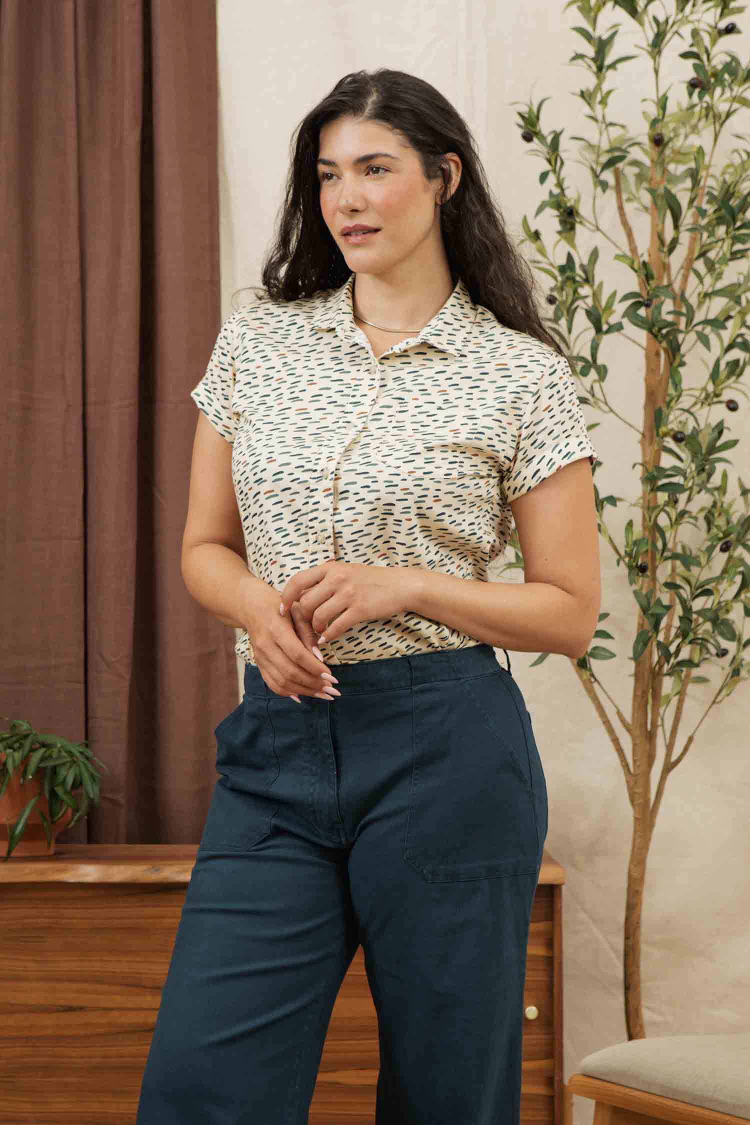A woman with long dark hair stands indoors wearing the Bridge & Burn Bea Shirt in Ivory Dashes, tucked into high-waisted navy pants. A wooden dresser, potted plant, and leafy tree are visible in the background.