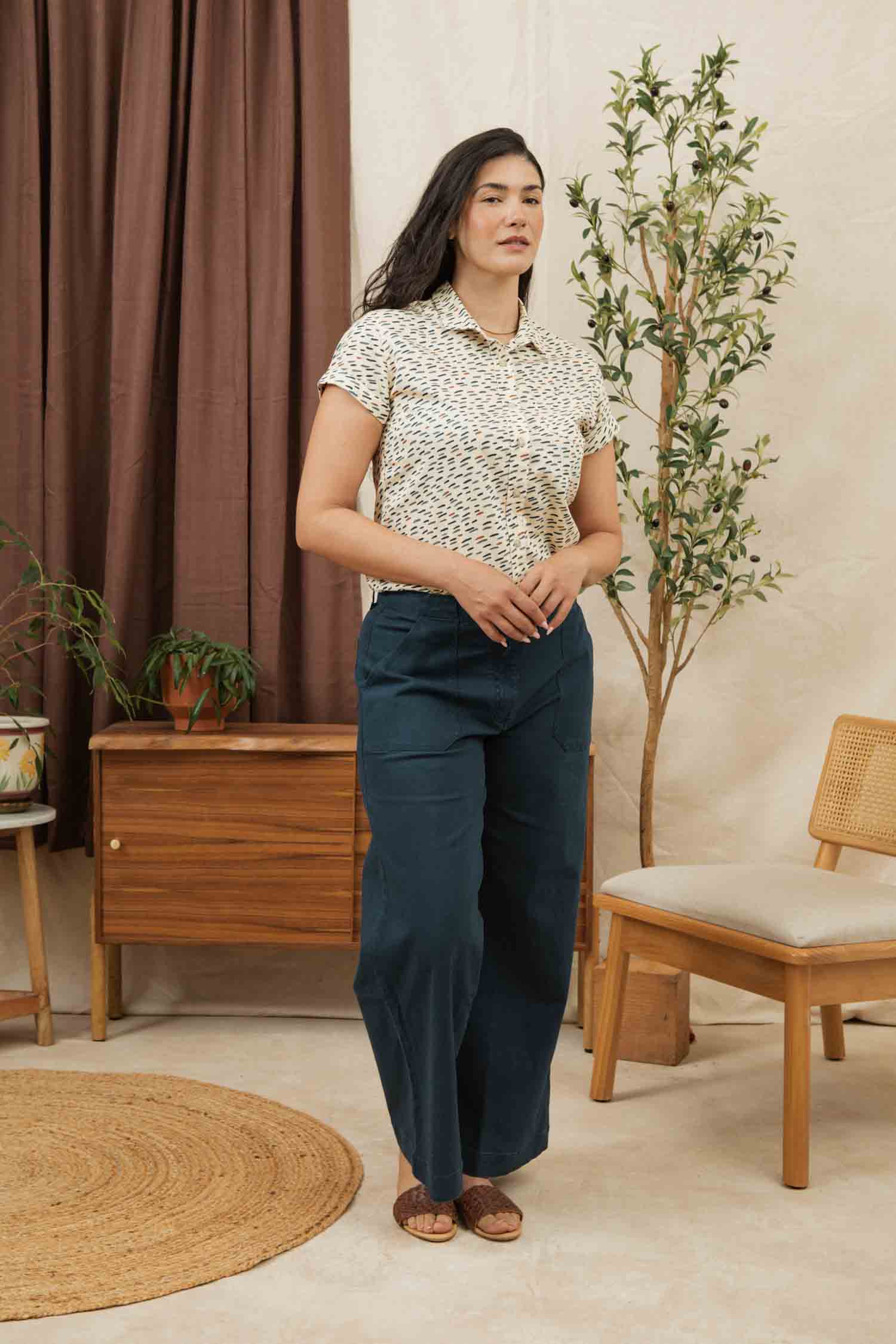 A woman stands indoors wearing the Bridge & Burn Bea Shirt in Ivory Dashes, styled with wide-leg blue pants. She is beside a wooden cabinet and chair, with potted plants and brown curtains in the background.
