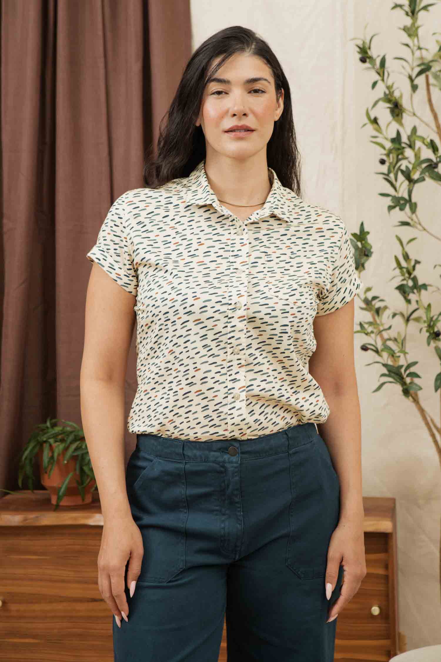 A woman with long dark hair stands indoors, wearing the Bridge & Burn Bea Shirt in Ivory Dashes and blue pants. Behind her are a wooden table with a potted plant, leafy greenery, and brown curtains for a cozy touch.