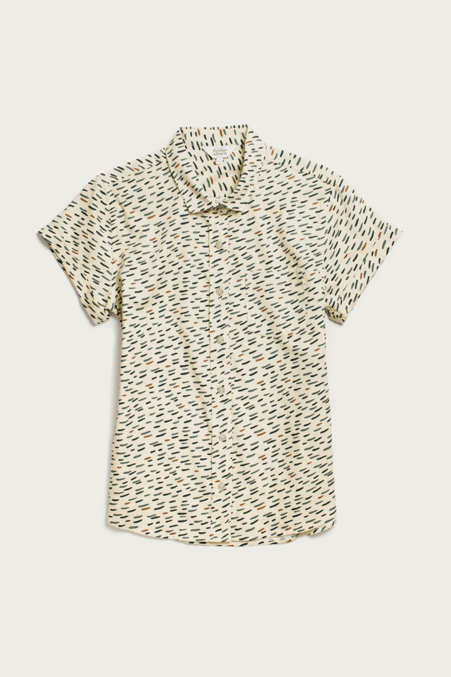 The Bridge & Burn Bea Shirt in Ivory Dashes is a classic short-sleeve button-down featuring a collar and scattered black, green, and brown dashes on a white background, shown laid flat on a white surface.