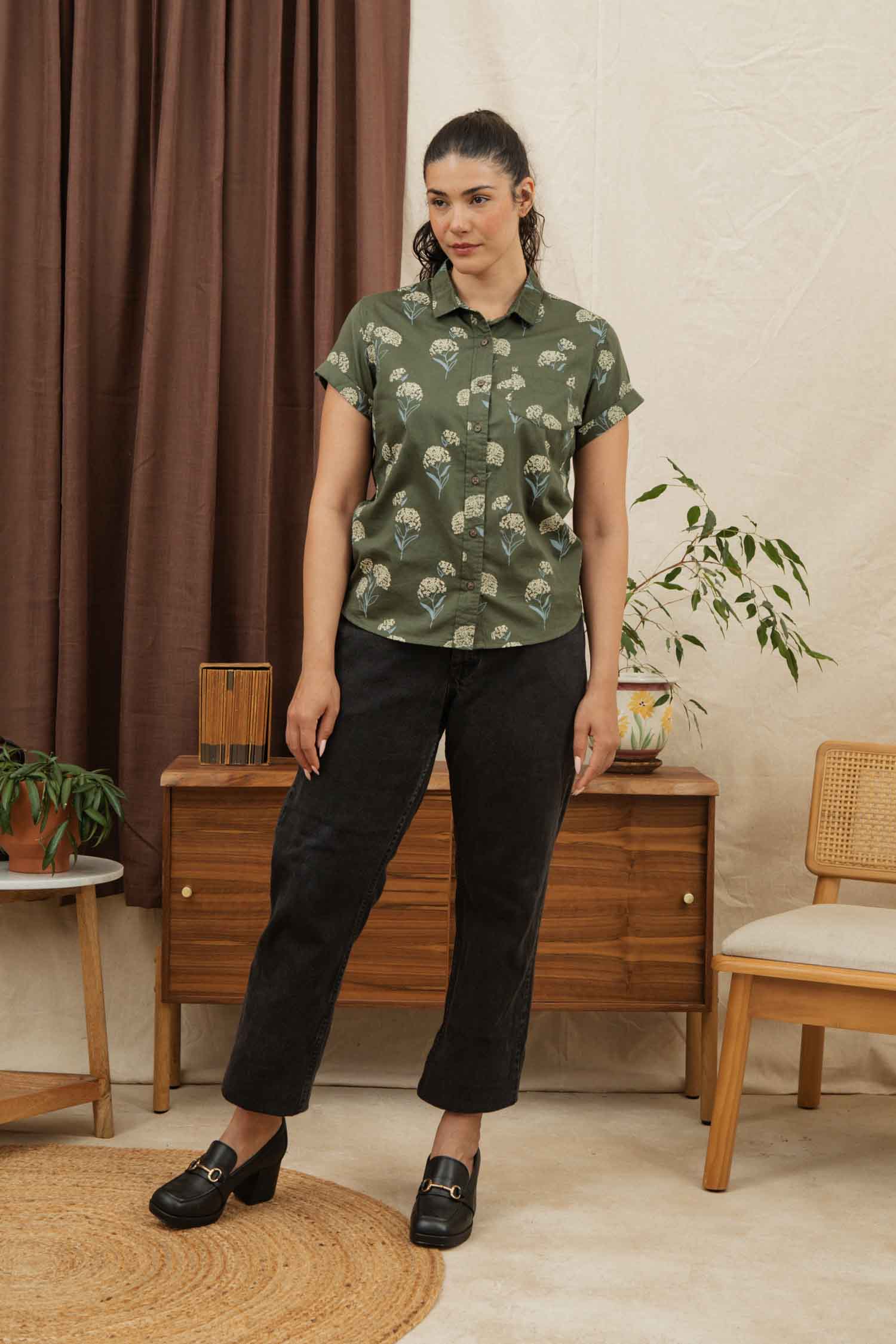 A woman with dark hair tied back wears the Bridge & Burn Bea Shirt in Olive Wildflower, featuring a green organic cotton short-sleeve button-down with a white pattern, paired with black pants. She stands indoors near a wooden cabinet and plants.