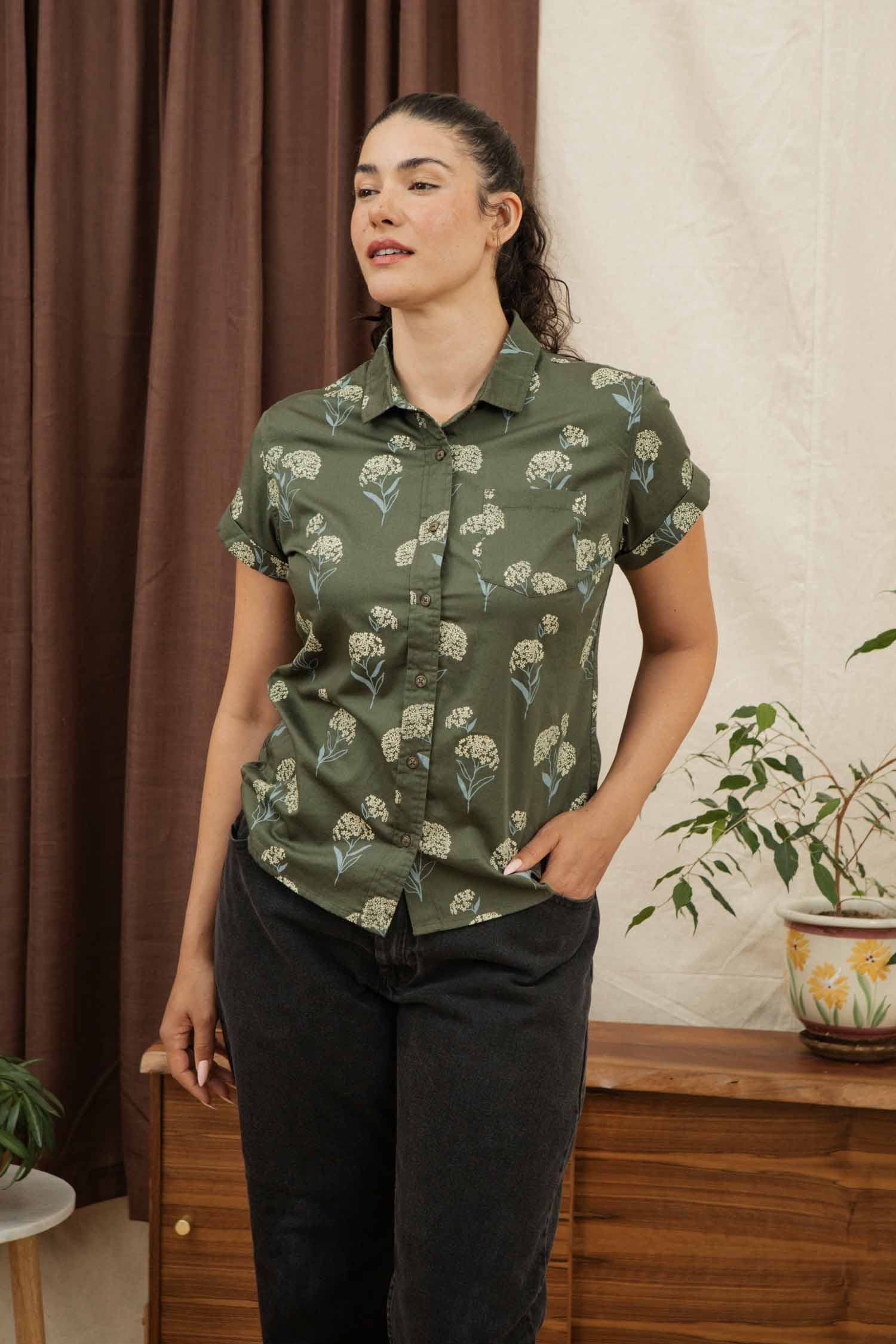 A woman with dark hair in a ponytail wears the Bridge & Burn Bea Shirt in Olive Wildflower and dark pants, standing indoors near a wooden table with a potted plant.
