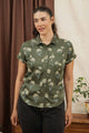 A woman with dark hair tied back wears the Bridge & Burn Bea Shirt in Olive Wildflower—a short-sleeve, tailored button-down with a floral print. She stands indoors by brown and cream curtains next to a small table with a potted plant.