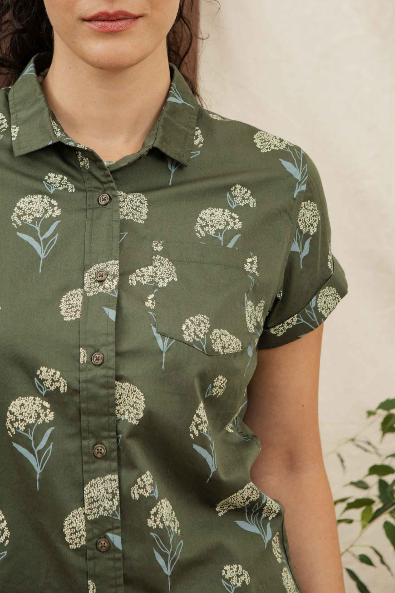 A woman models the Bridge & Burn Bea Shirt in Olive Wildflower, a tailored fit short-sleeve button down with a white floral print, shown from shoulders to waist against a light background with a small leafy plant.