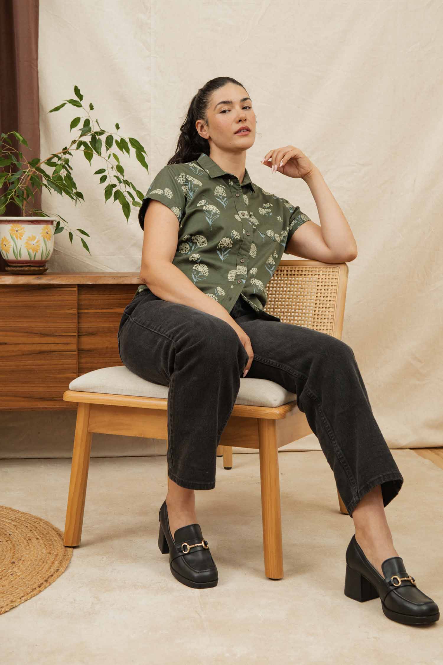 A woman with dark hair sits on a wooden chair wearing the Bridge & Burn Bea Shirt in Olive Wildflower, styled with black jeans and black heeled loafers. A potted plant on a sideboard enhances the cozy, modern setting.