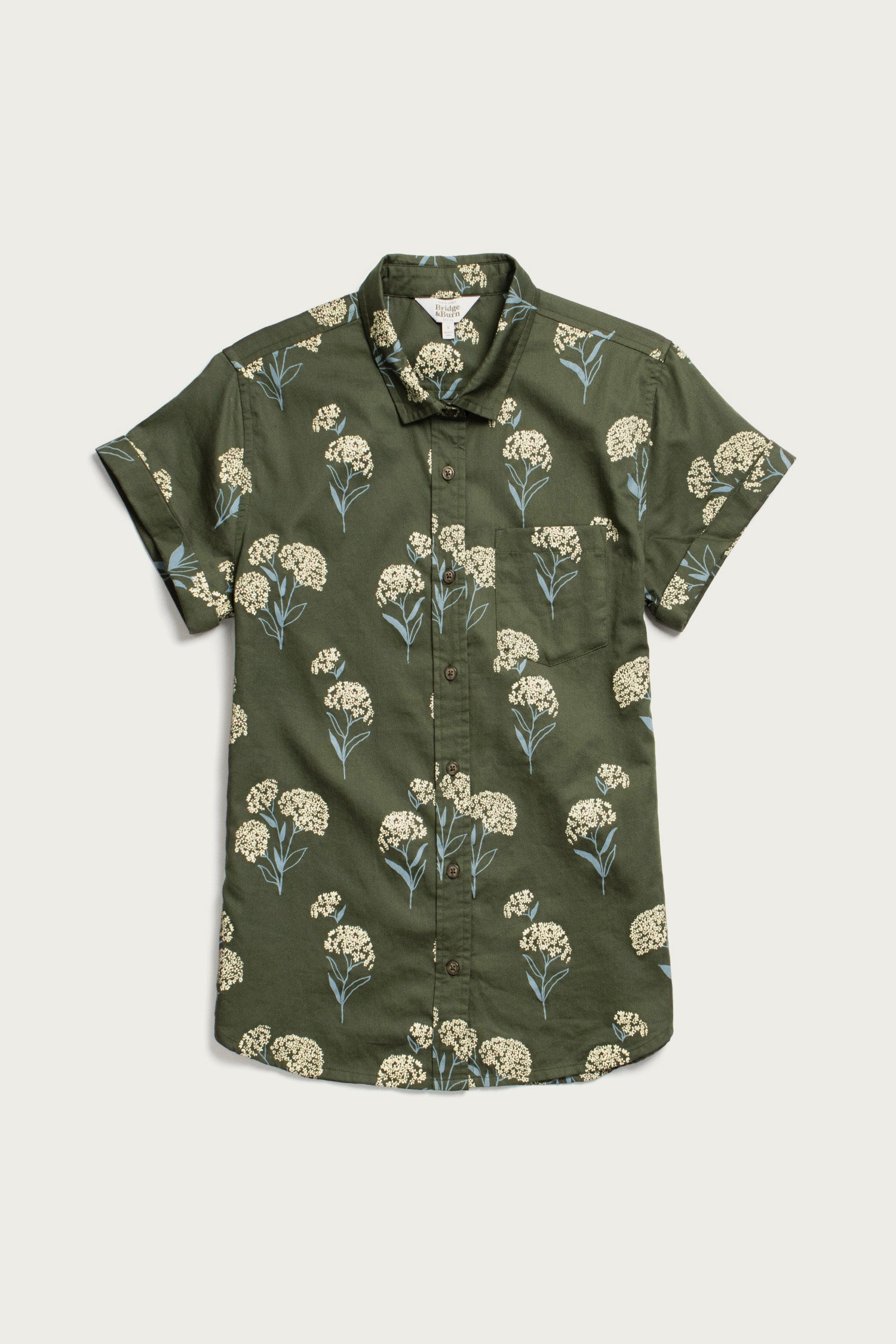 The Bridge & Burn Bea Shirt in Olive Wildflower is an organic cotton, short-sleeve button-down with a tailored fit, collar, chest pocket, and an olive green base featuring white and blue floral patterns with green stems.