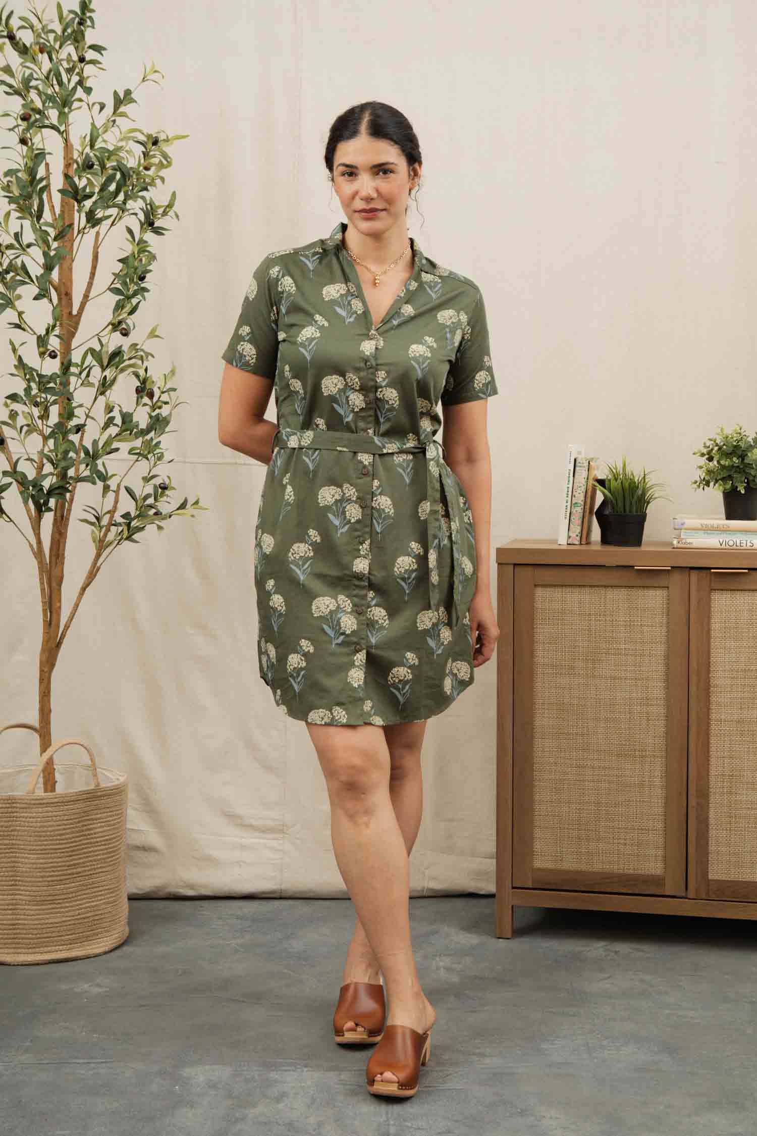 A woman stands indoors near a plant and wooden cabinet, wearing the Bridge & Burn Sofia Shirt Dress in Olive Wildflower—an organic cotton dress with a waist tie—paired with brown open-toe clogs and her hair tied back.