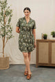 A woman stands indoors near a plant and wooden cabinet, wearing the Bridge & Burn Sofia Shirt Dress in Olive Wildflower—an organic cotton dress with a waist tie—paired with brown open-toe clogs and her hair tied back.