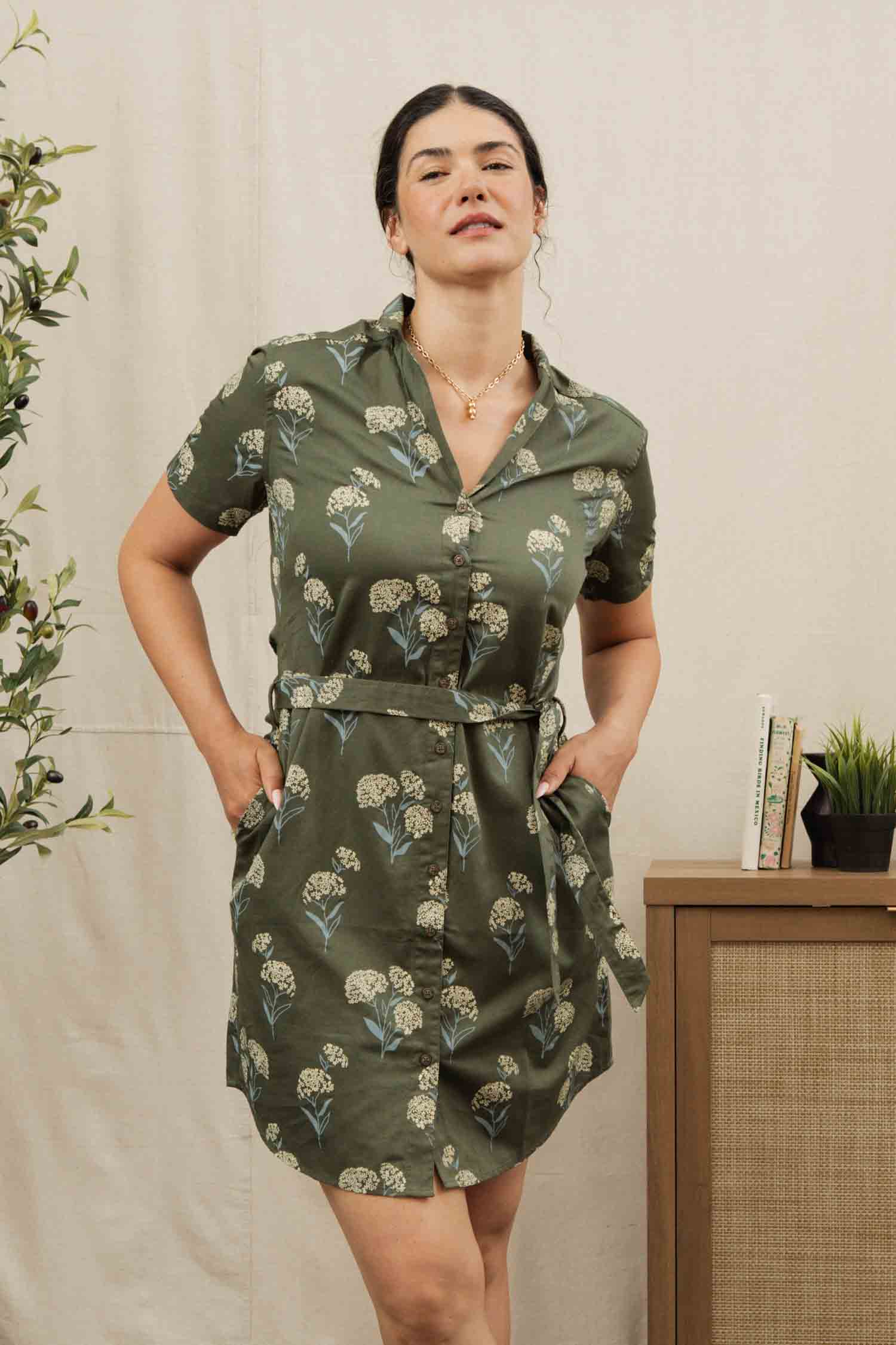 A woman wears the Bridge & Burn Sofia Shirt Dress in Olive Wildflower—a short-sleeved, belted, floral button-front style with pockets. A plant and a cabinet with books and a potted plant appear in the background.