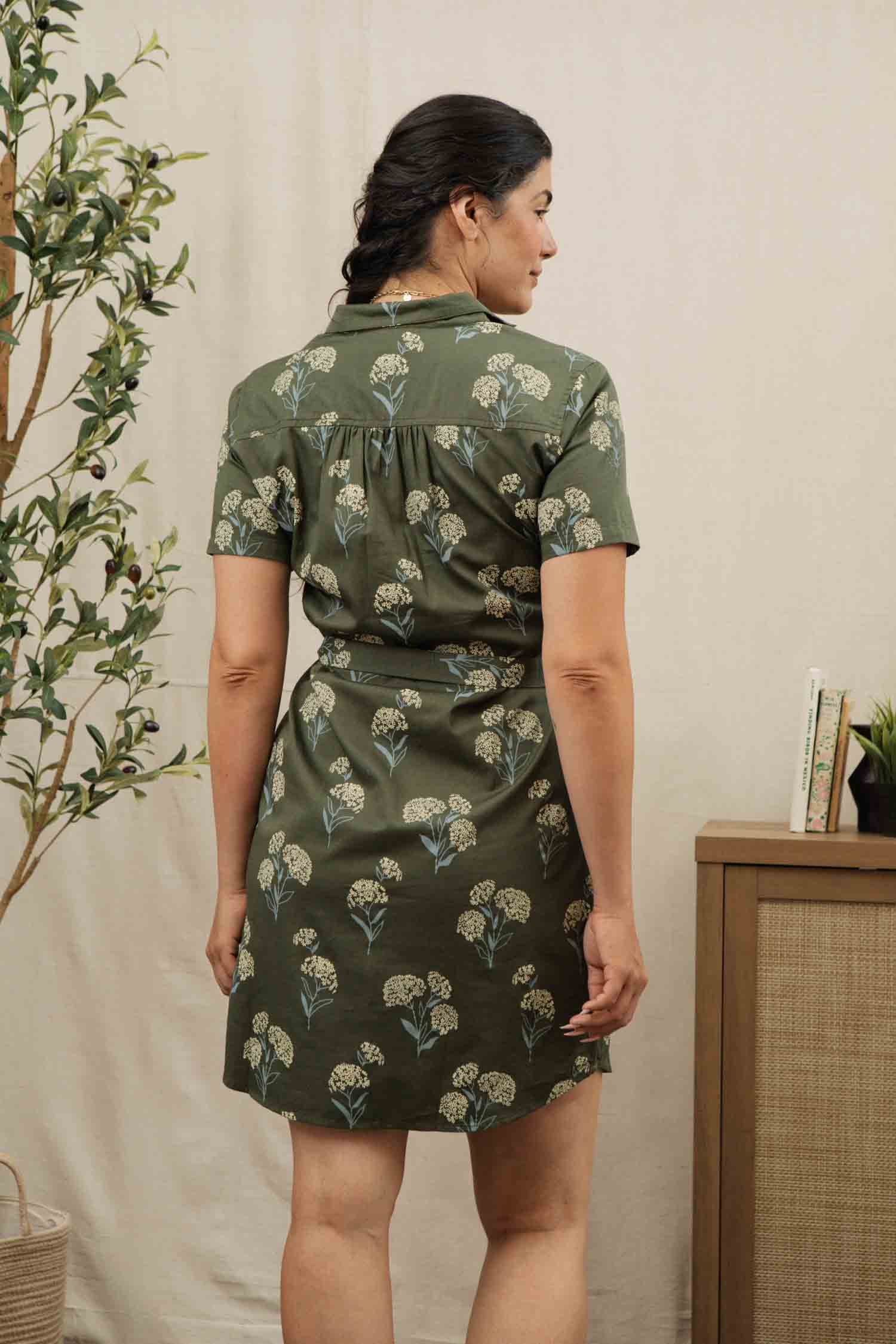 A woman with dark hair in a low braid wears the Bridge & Burn Sofia Shirt Dress in Olive Wildflower, standing indoors with her back to the camera near a wooden cabinet and a leafy potted plant.