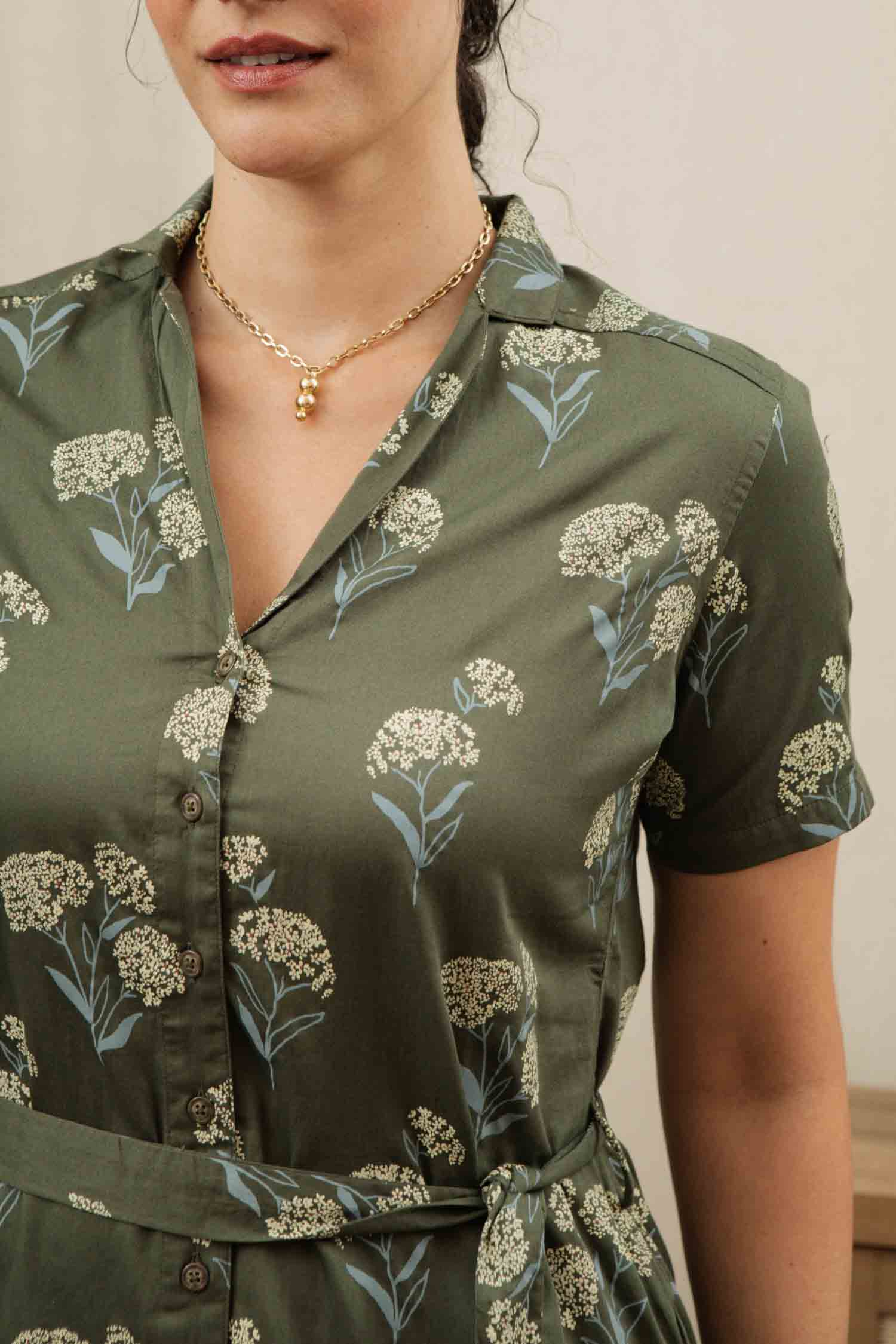 A woman wears the Bridge & Burn Sofia Shirt Dress in Olive Wildflower, featuring short sleeves and a white floral print. She also wears a gold necklace. The photo is cropped to show her upper body without revealing her full face.