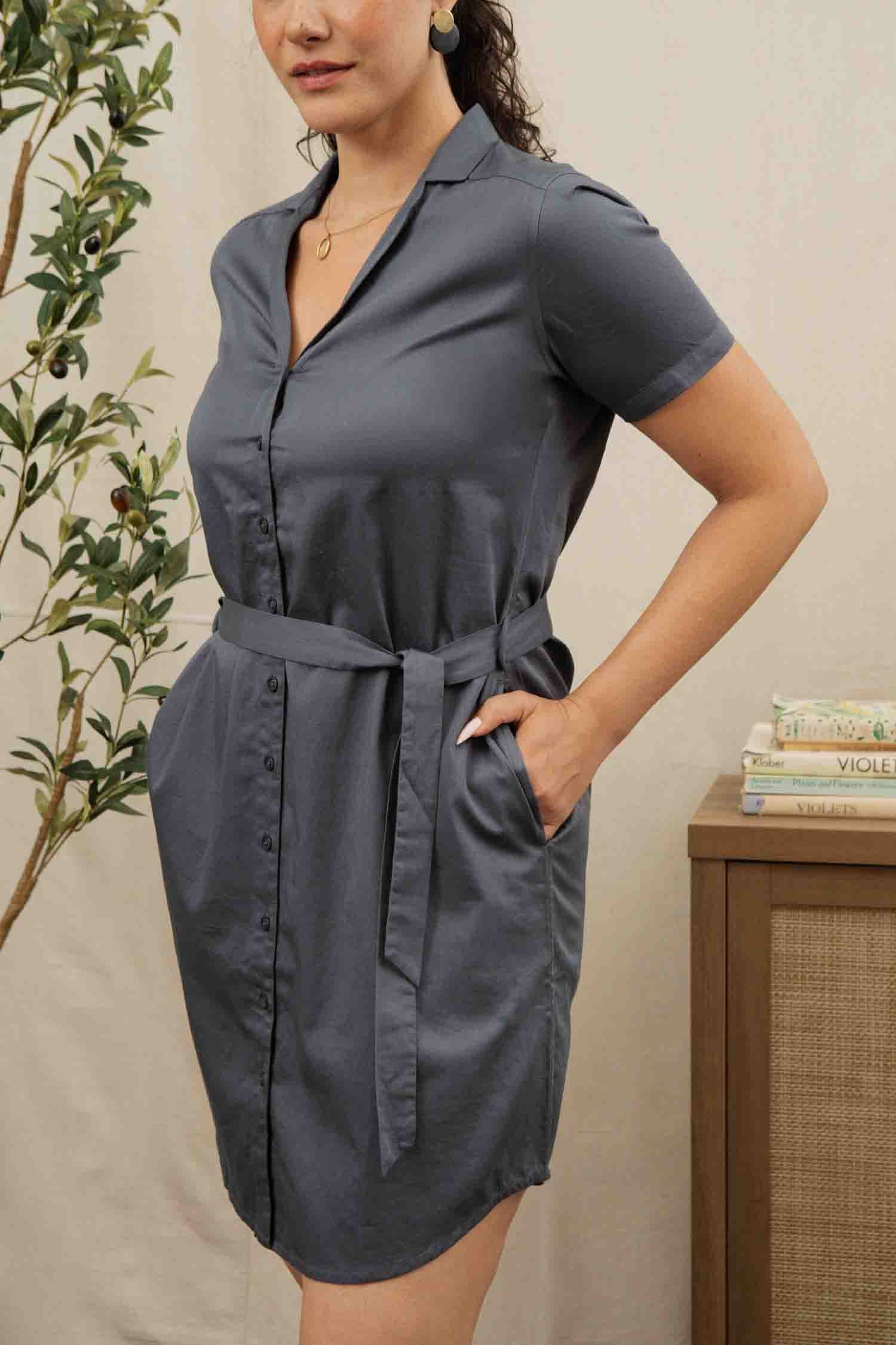 A woman wears the Bridge & Burn Sofia Shirt Dress in light navy—short-sleeved, button-front with a belt and pockets—standing indoors beside a leafy plant and a small table stacked with books.