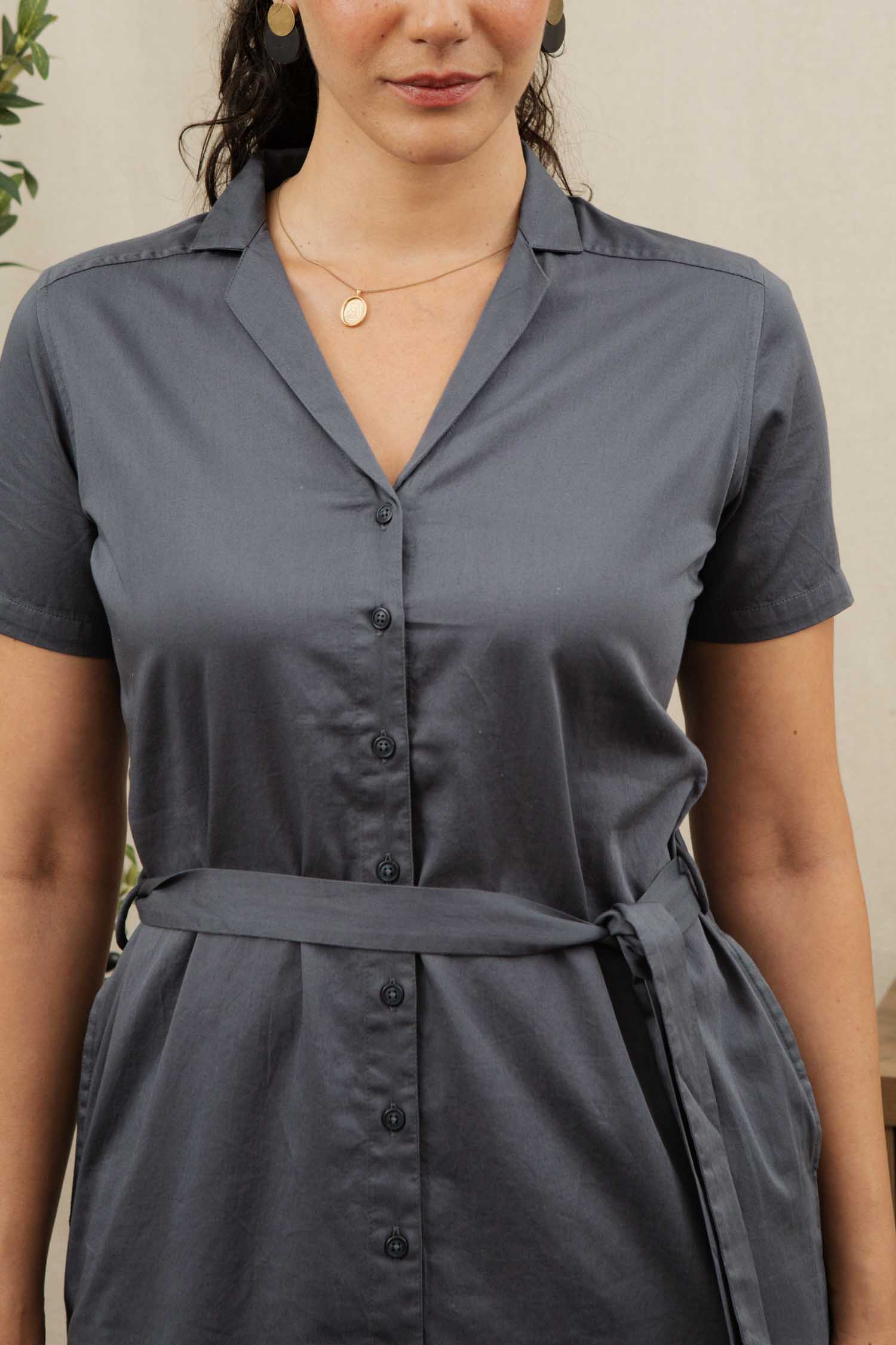 A woman stands indoors wearing the Bridge & Burn Sofia Shirt Dress in Light Navy, featuring a button front and waist tie. She accessorizes with gold earrings and a necklace, her curly hair resting on her shoulder, face partially visible.