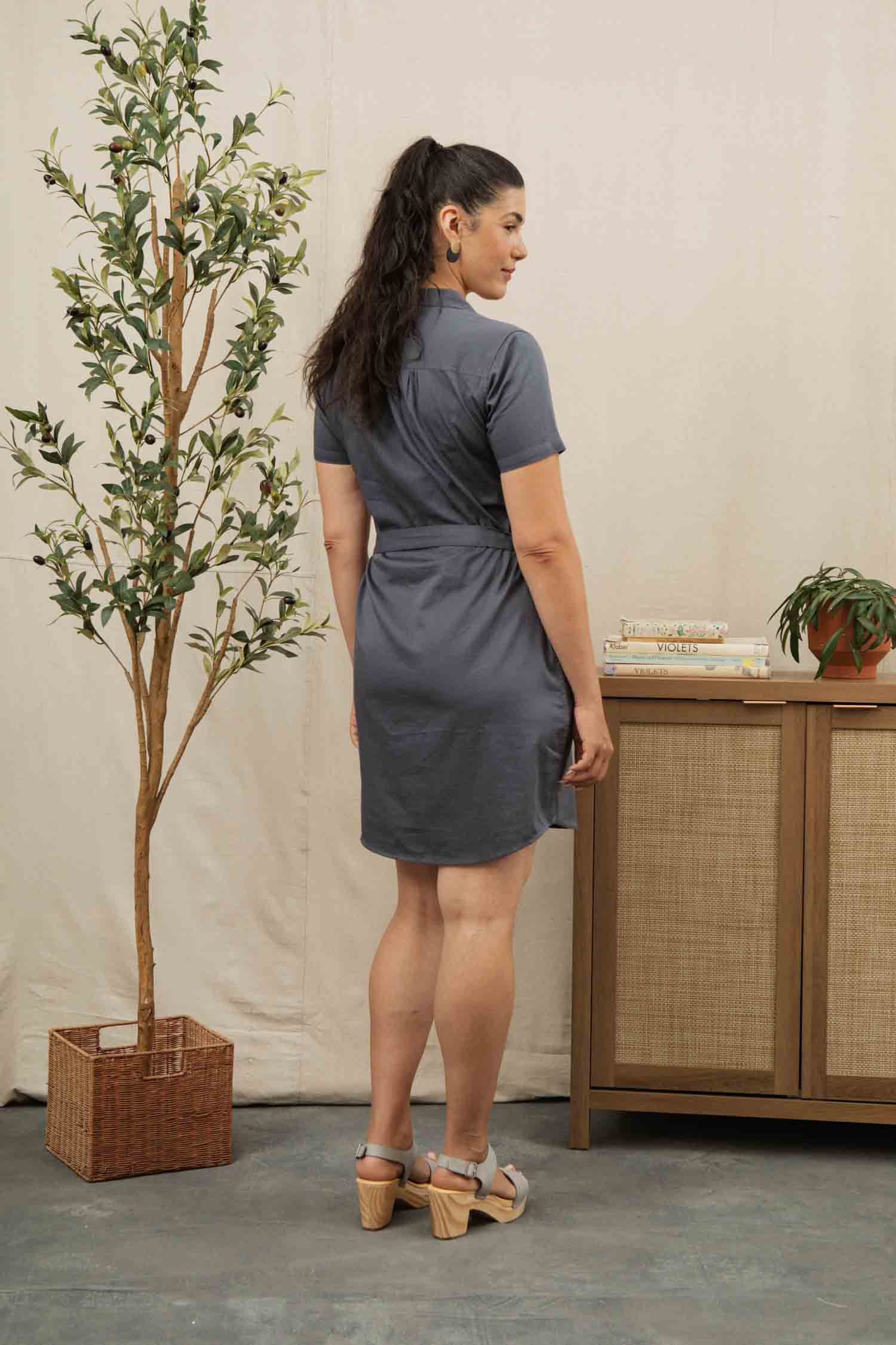 A woman with dark hair in a ponytail faces away, wearing the Bridge & Burn Sofia Shirt Dress in Light Navy and wooden-heeled sandals. She stands near a small tree and a wooden sideboard decorated with books and a potted plant.