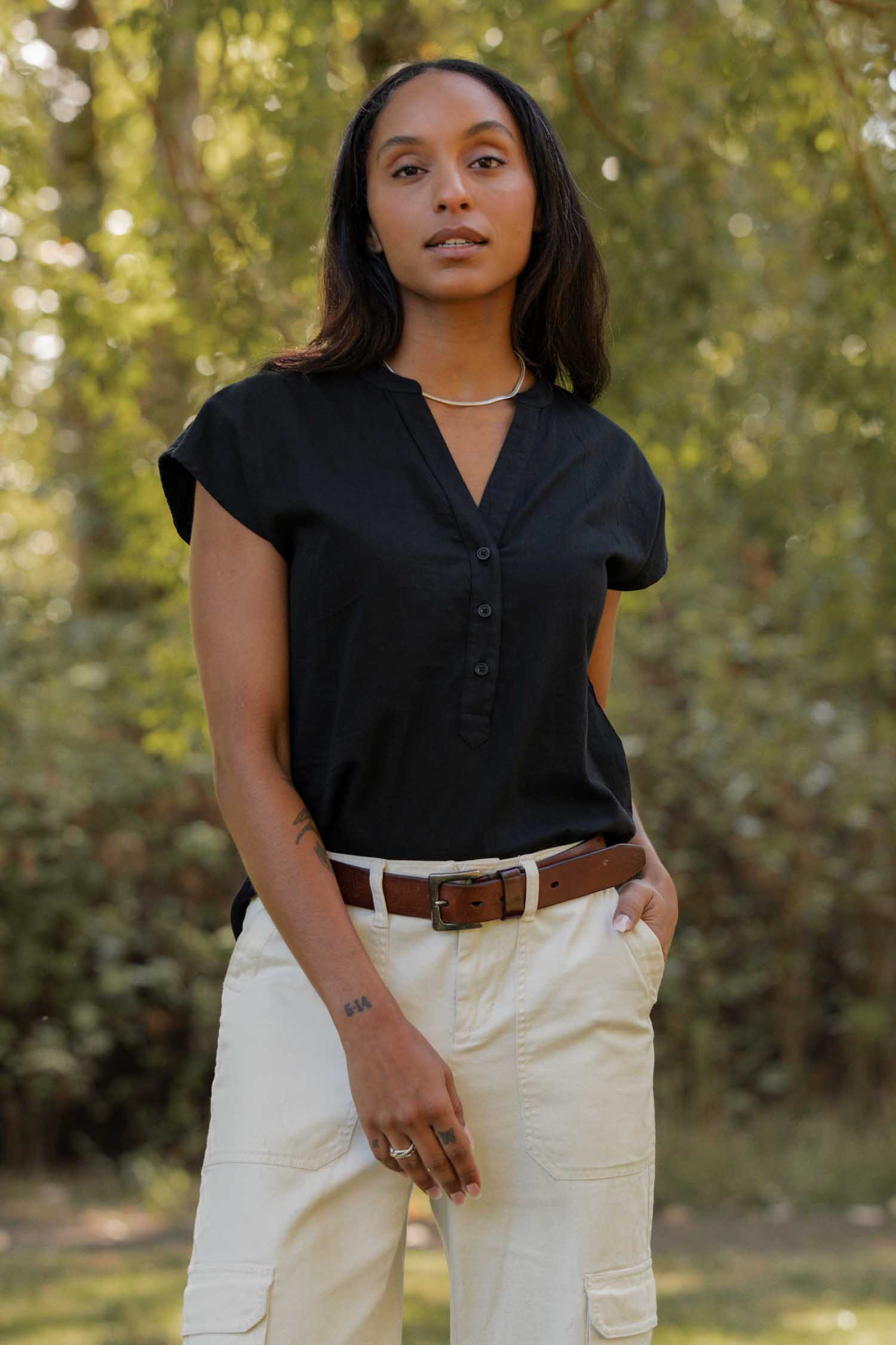 A person stands outdoors in front of green foliage, wearing the Bridge & Burn Ladd Shirt in black, tucked into beige pants with a brown belt, looking confidently at the camera.