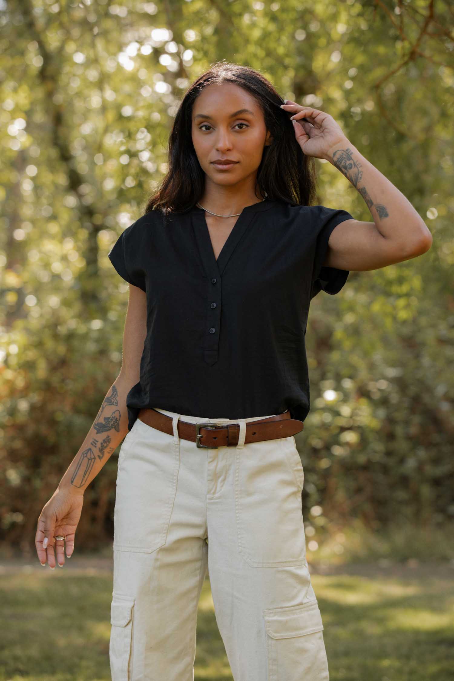 A woman with long dark hair and tattoos stands outdoors among green foliage, wearing the Bridge & Burn Ladd Shirt in black, tucked into beige cargo pants with a brown belt.