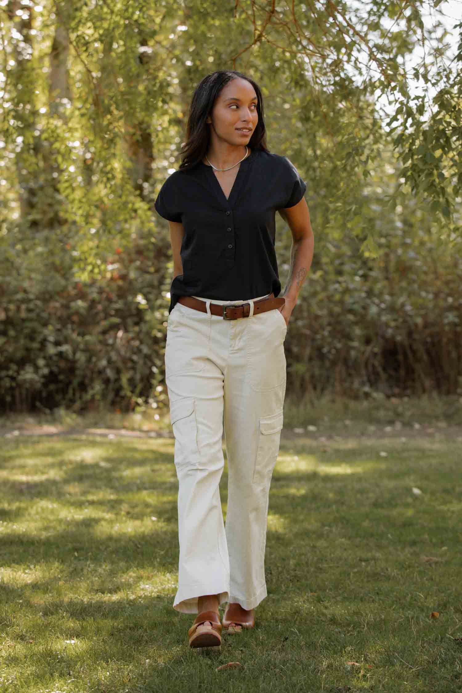 Wearing the Bridge & Burn Ladd Shirt in black, a woman pairs it with white cargo pants and sandals as she walks on grass in a sunlit park, surrounded by trees and greenery.