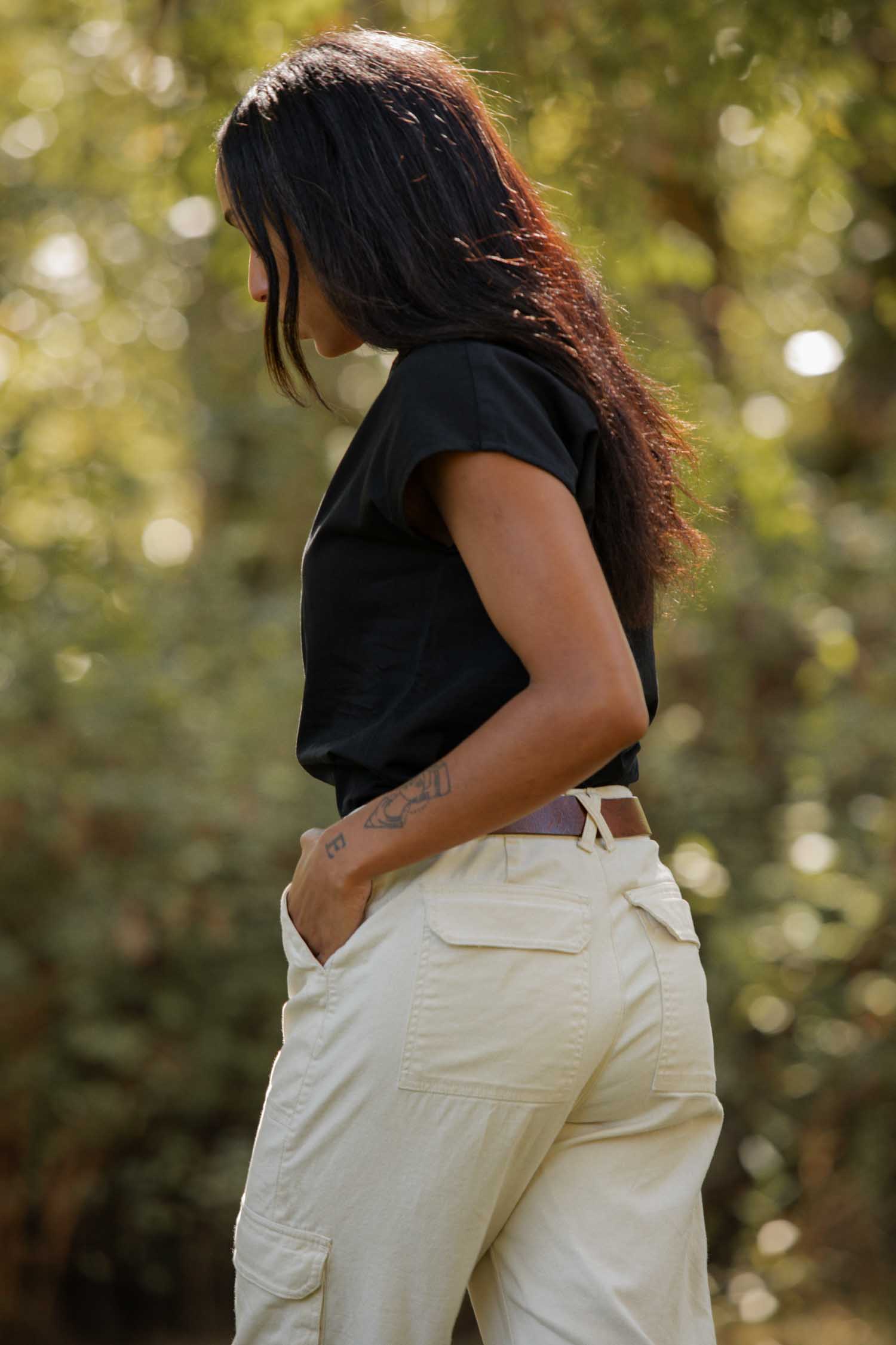 A woman with long dark hair wears the Bridge & Burn Ladd Shirt in black and light-colored pants, standing outdoors in profile with her hand in her pocket as sunlight streams through green foliage behind her.