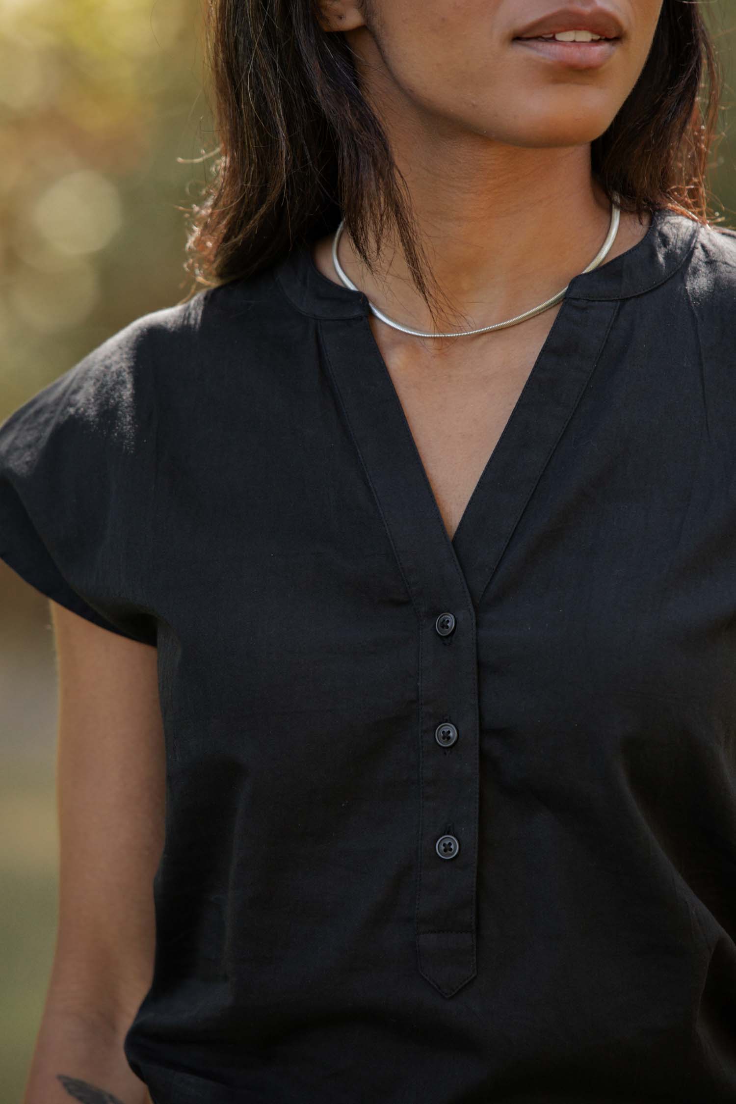 A person wearing the Bridge & Burn Ladd Shirt in black, a short sleeve button-up, paired with a silver necklace, stands outdoors with blurred greenery in the background.