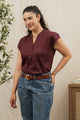 A woman with long dark hair in a ponytail wears the Bridge & Burn Ladd Shirt in burgundy, paired with blue jeans and a brown belt. Indoors by a leafy plant and stacked books, she smiles slightly—her look blends comfort and feminine style.