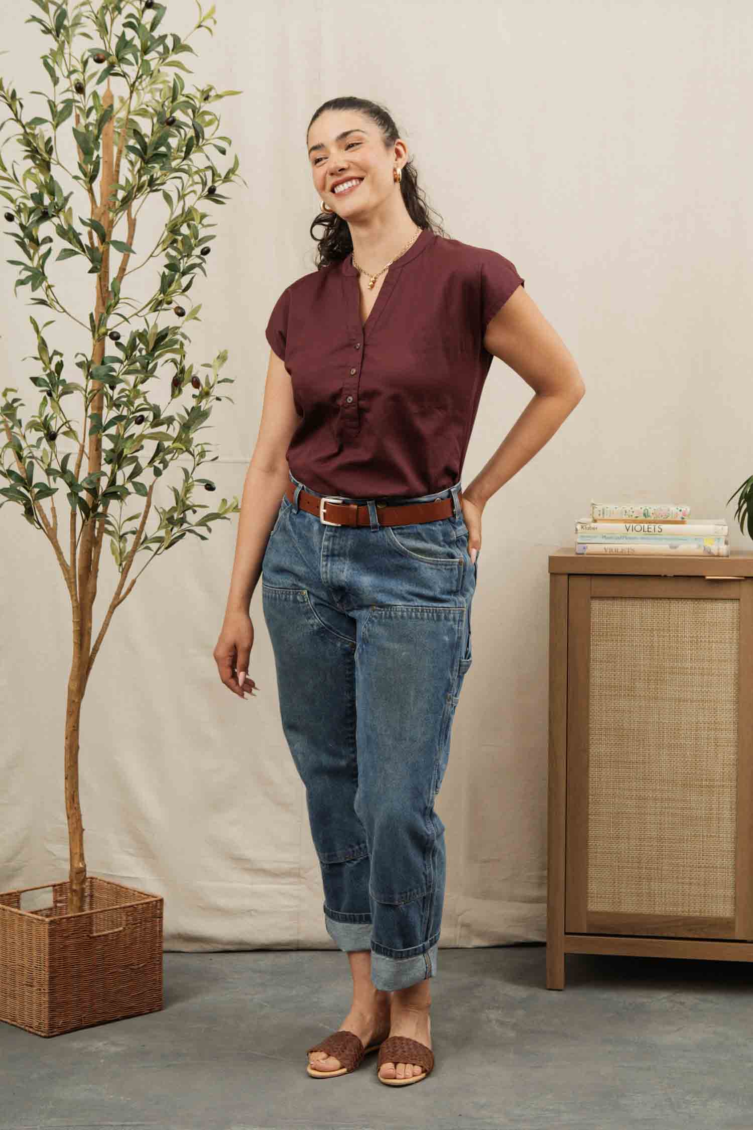 A woman wears the Bridge & Burn Ladd Shirt in burgundy, styled with rolled-up blue jeans and brown sandals, smiling with one hand in her pocket next to a potted plant and a cabinet topped with books.