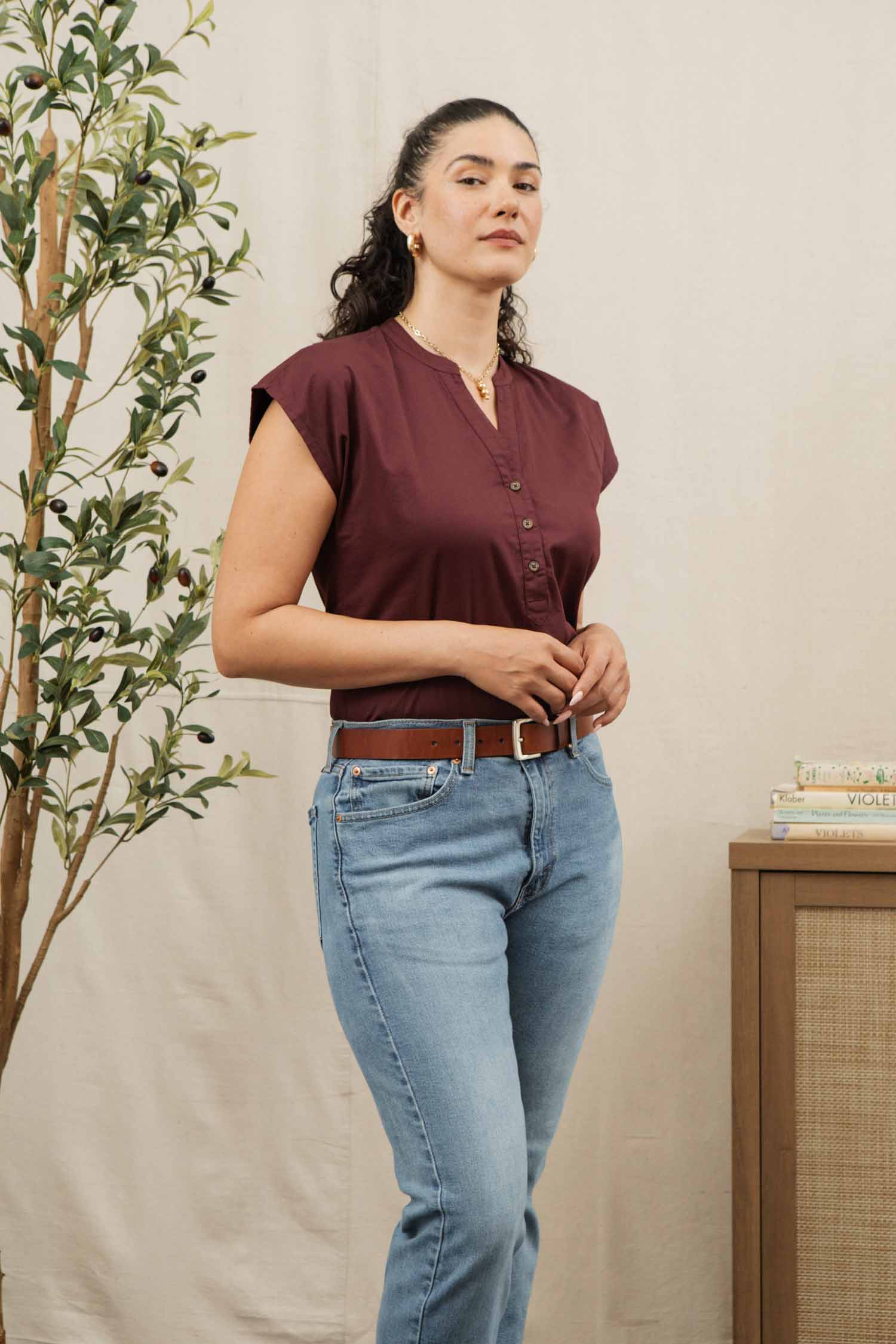 A woman with dark, curly hair wears the Bridge & Burn Ladd Shirt in burgundy and blue jeans. Standing by a leafy plant and a table of books, she looks confidently at the camera.