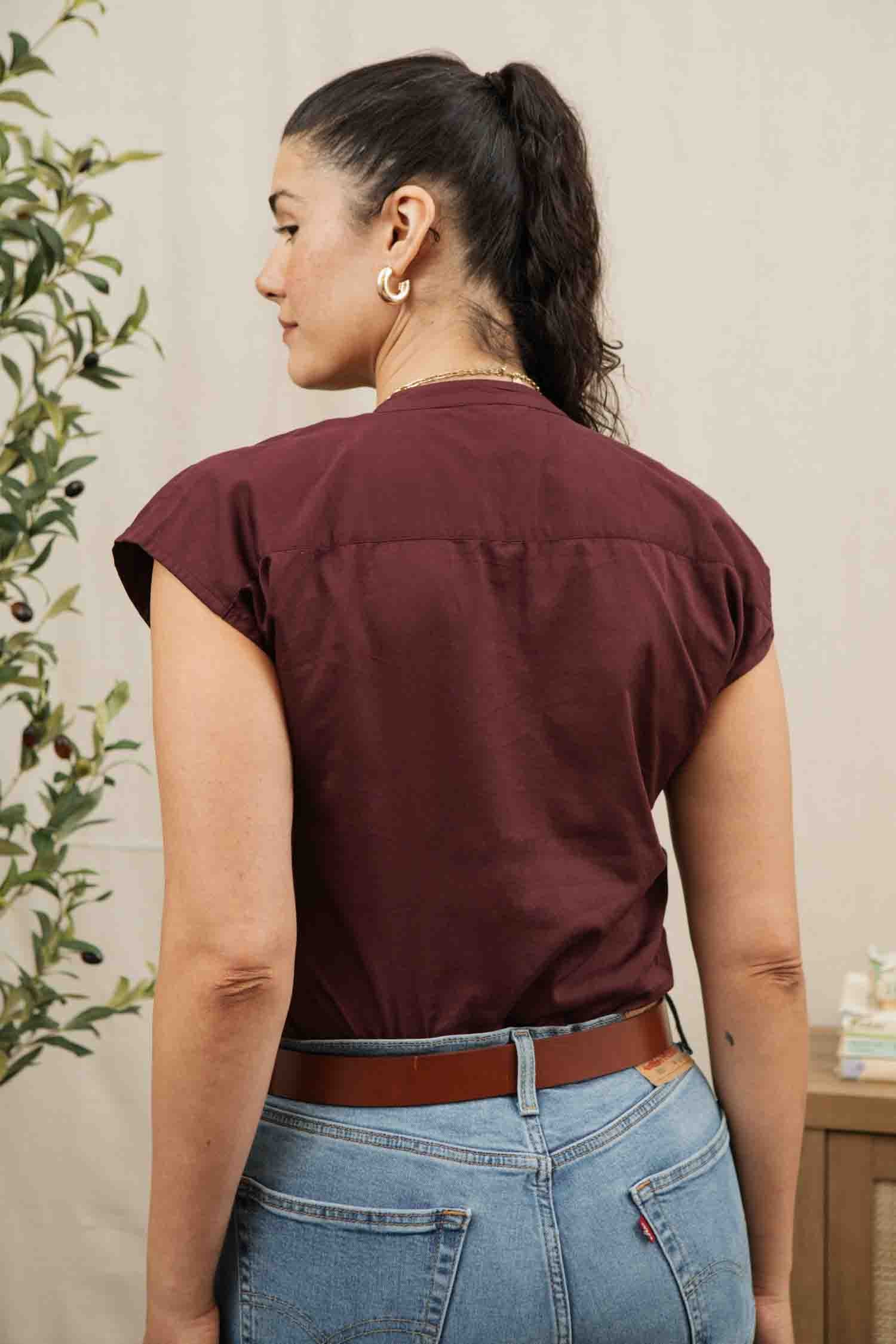 A woman with dark hair in a ponytail stands with her back to the camera, wearing the Bridge & Burn Ladd Shirt in burgundy and blue jeans. A plant is on the left, and a small wooden table with items is on the right.