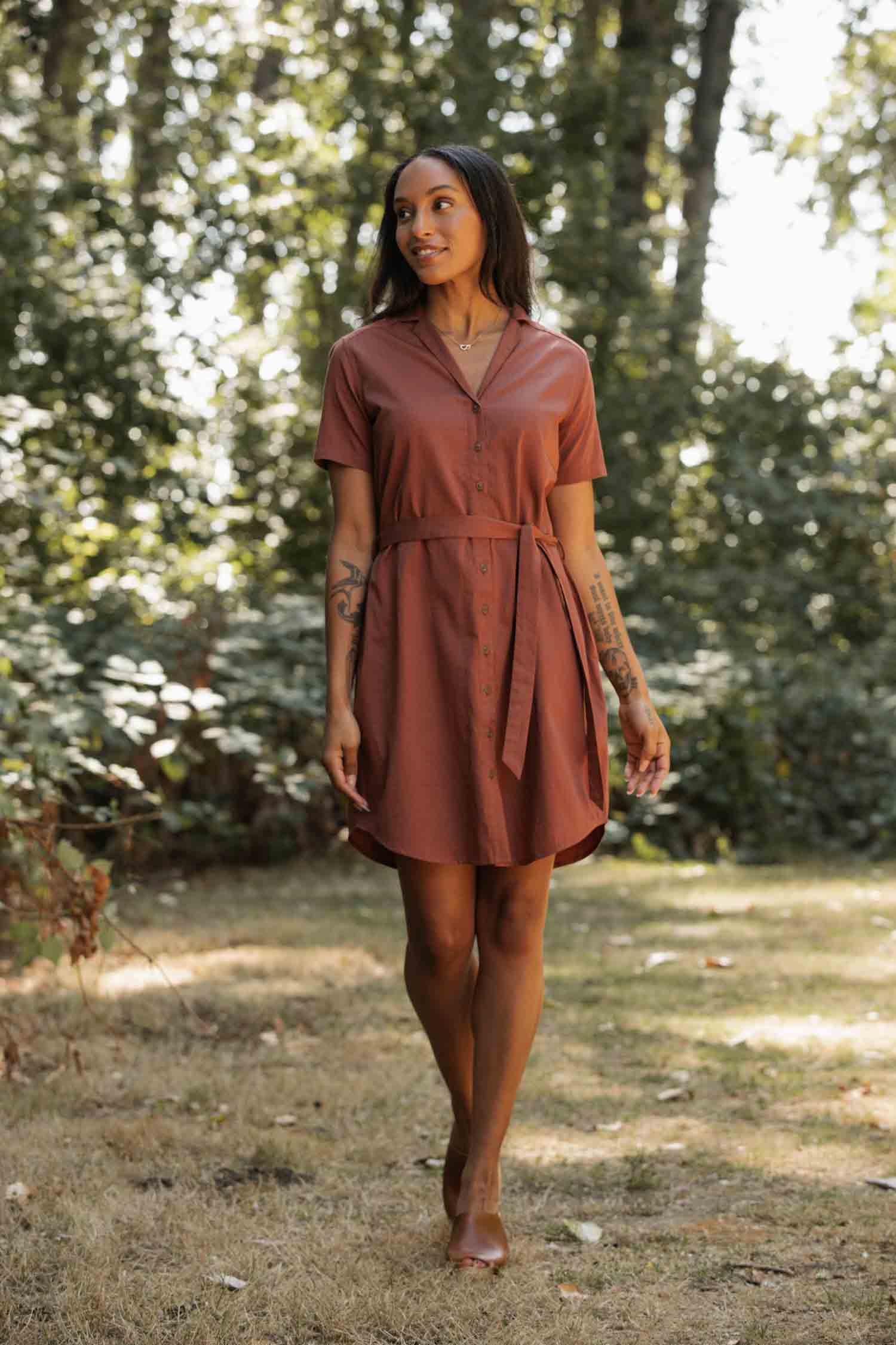 A woman wearing the Bridge & Burn Sofia Shirt Dress in Copper walks on grass in a sunlit, wooded area, looking to the side and smiling softly.