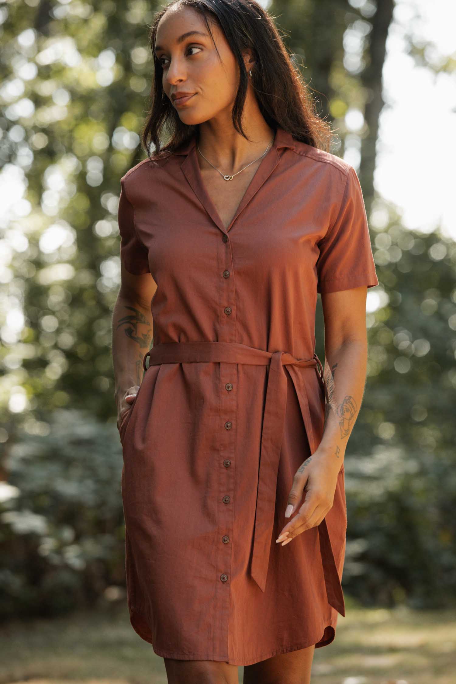 A woman stands outdoors with her hands in her pockets, wearing the Bridge & Burn Sofia Shirt Dress in Copper, a short-sleeved, button-front style with a waist tie, surrounded by lush greenery and dappled sunlight.