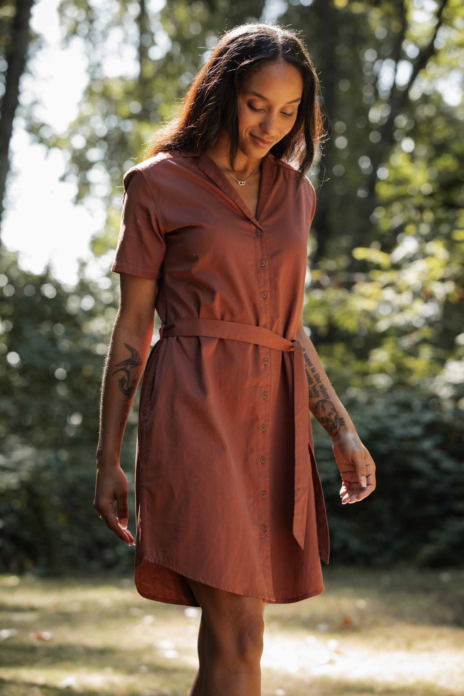 A woman stands outdoors among green trees and sunlight, smiling softly while wearing the Bridge & Burn Sofia Shirt Dress in Copper.