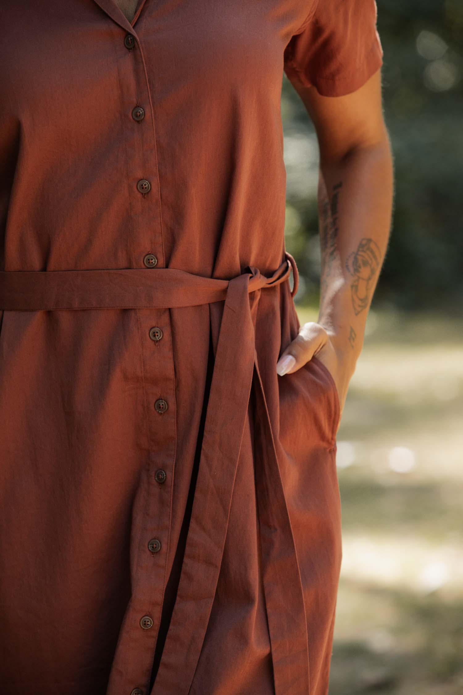 A person stands outdoors in the Bridge & Burn Sofia Shirt Dress in Copper, an organic cotton piece with a button front and fabric belt. One hand is in the pocket, revealing a forearm tattoo, set against blurred greenery.