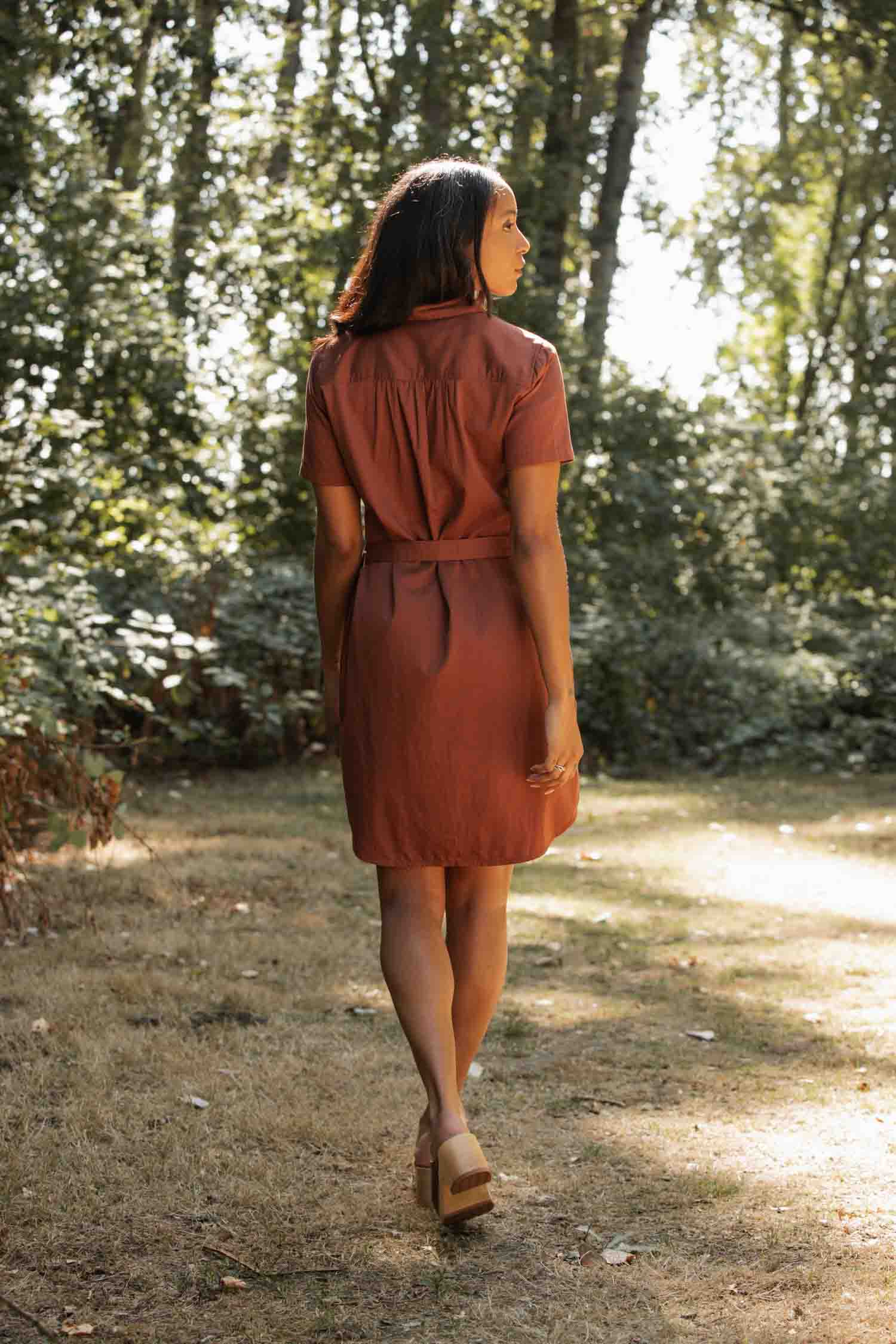A woman in the Bridge & Burn Sofia Shirt Dress in Copper and sandals walks away on a sunlit path surrounded by trees and greenery.
