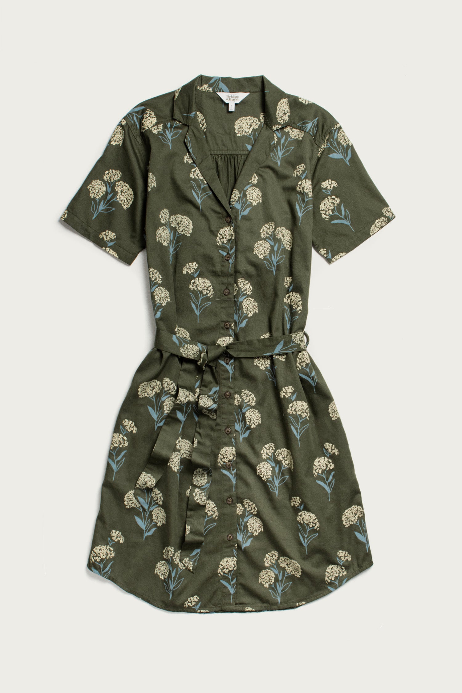 The Sofia Shirt Dress by Bridge & Burn is a short-sleeve, button-front dress in olive green organic cotton, featuring a belted waist and a white and blue wildflower print with green leaves. The dress is shown flat against a plain background.