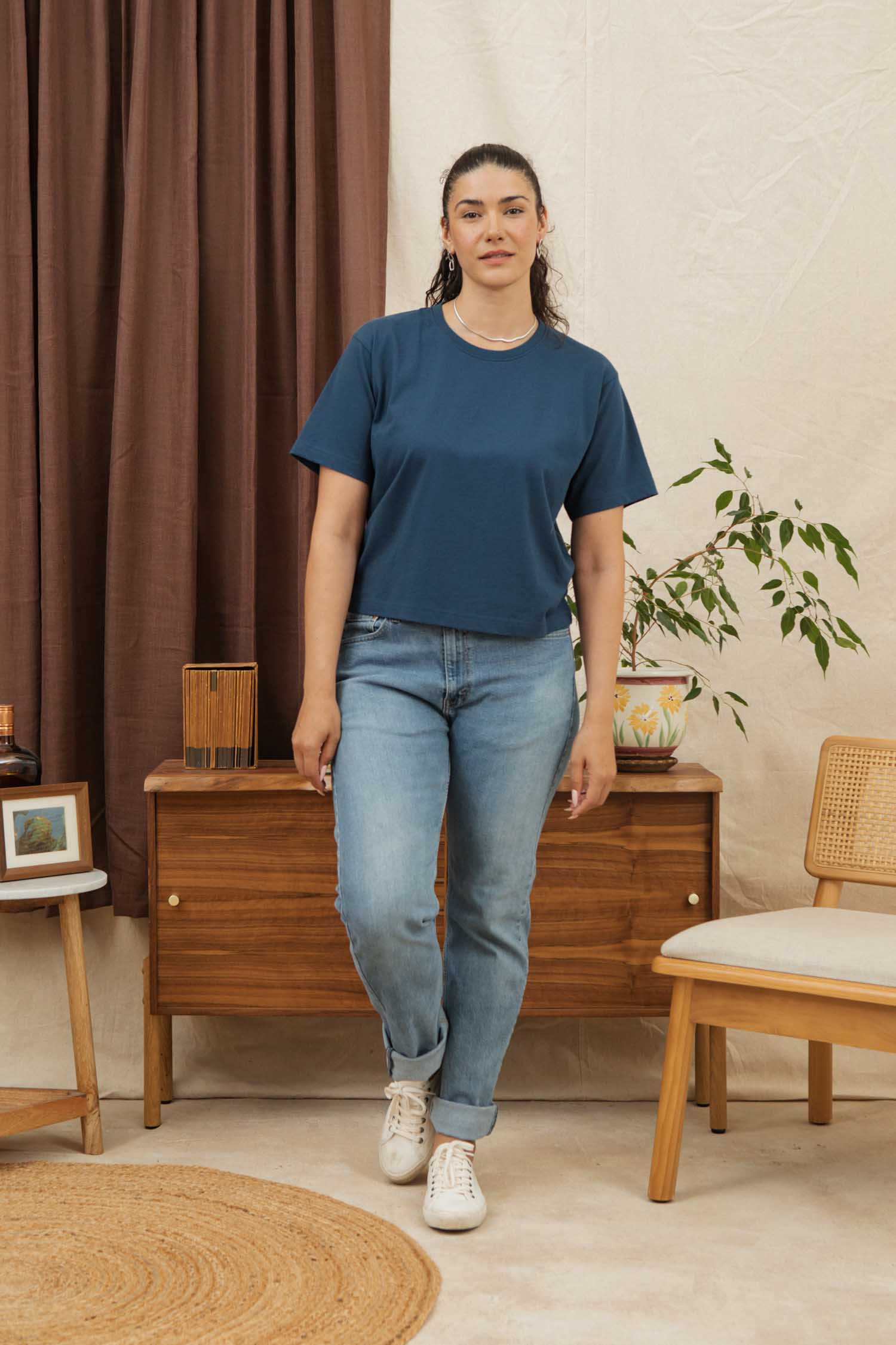 A woman with dark hair in a ponytail stands indoors wearing the Bridge & Burn Recycled Boxy Tee in Cobalt, paired with light blue jeans and white sneakers. She is by a wooden console with plants, brown curtains, and a beige wall behind her.
