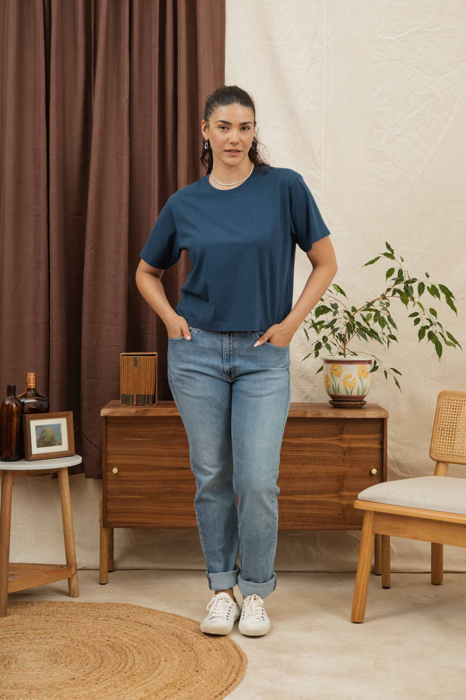 A woman with long dark hair tied back stands indoors wearing the Bridge & Burn Recycled Boxy Tee in Cobalt, light blue jeans, and white sneakers. She’s in front of a wooden cabinet with a plant and neutral decor around her.