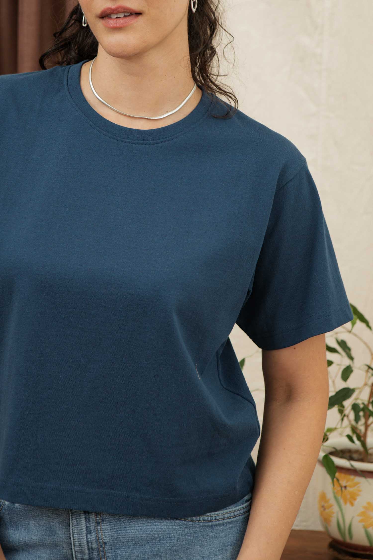 A woman stands indoors next to a potted plant, wearing the Bridge & Burn Recycled Boxy Tee in Cobalt and a silver necklace. Only her upper torso and part of her face are visible.