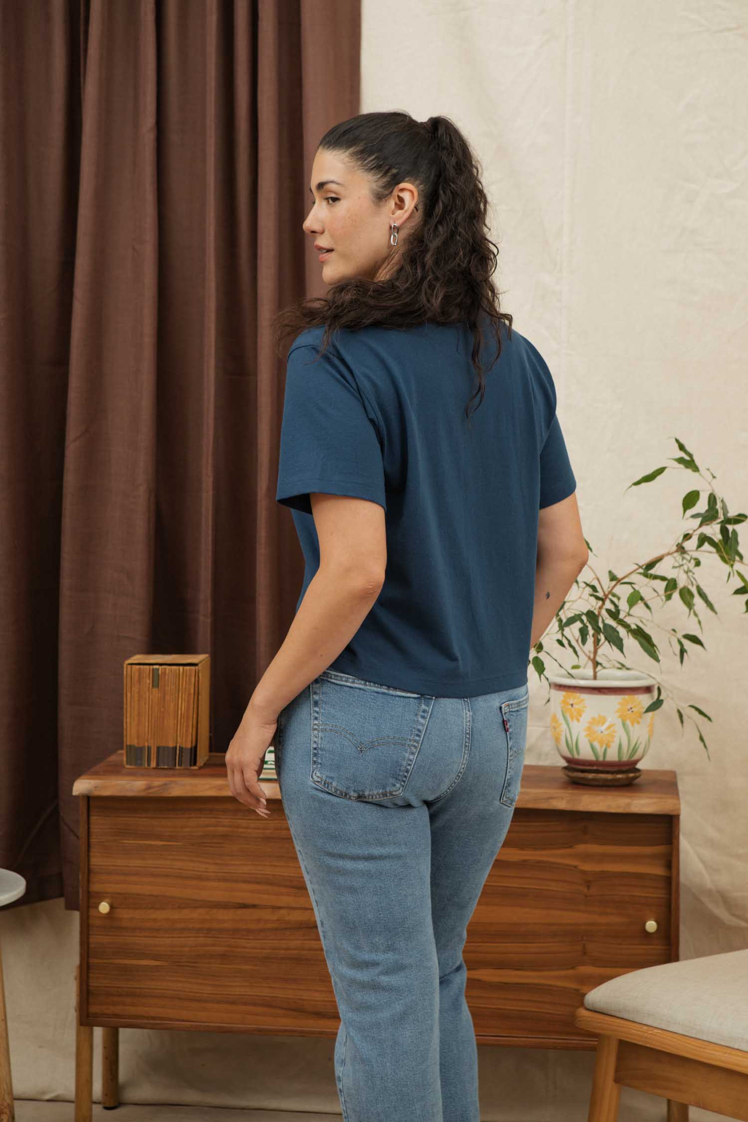 A woman with long, curly dark hair stands indoors in front of a wooden sideboard and potted plant, wearing Bridge & Burn’s Recycled Boxy Tee in Cobalt and light blue jeans. Brown curtains are visible behind her as she looks to the side.