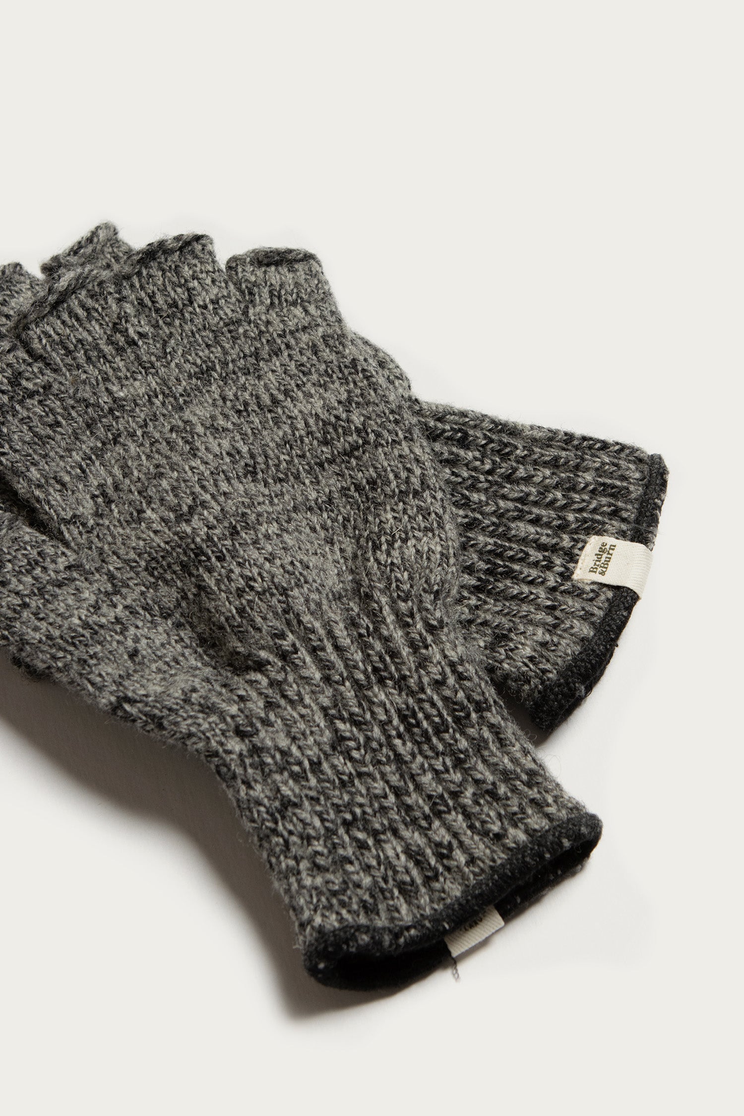 The Bridge & Burn Ragg Wool Fingerless Glove in Charcoal features a ribbed cuff and a small fabric tag, highlighting its cozy, practical design. The gray wool gloves are shown on a white background.