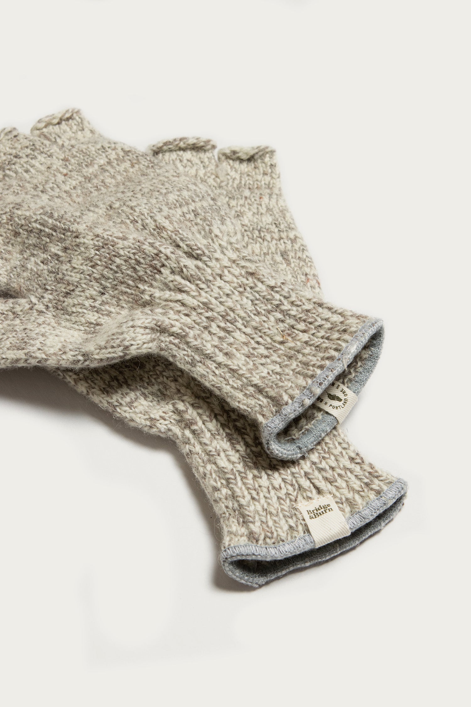Bridge & Burn's Ragg Wool Fingerless Glove in Oatmeal, featuring a visible woven texture and attached cuff tags, is shown in beige and gray tones resting on a white surface.