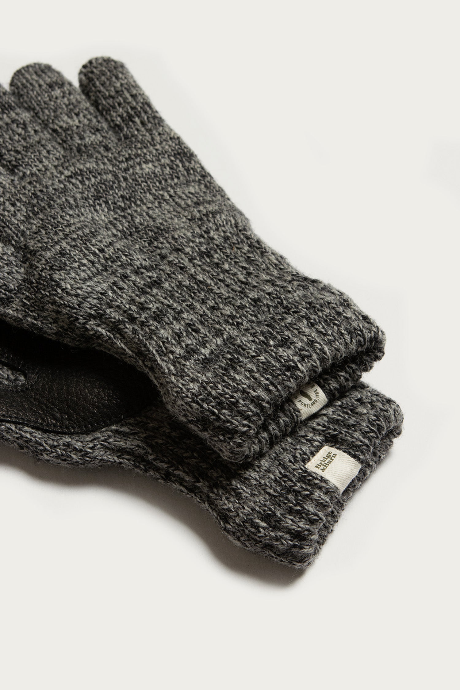 A close-up of Bridge & Burn’s Ragg Wool Lined Glove in Charcoal, stacked together on a plain background. The thick knit gloves have a subtle pattern, fabric tag on the cuff, and are proudly made in the USA.