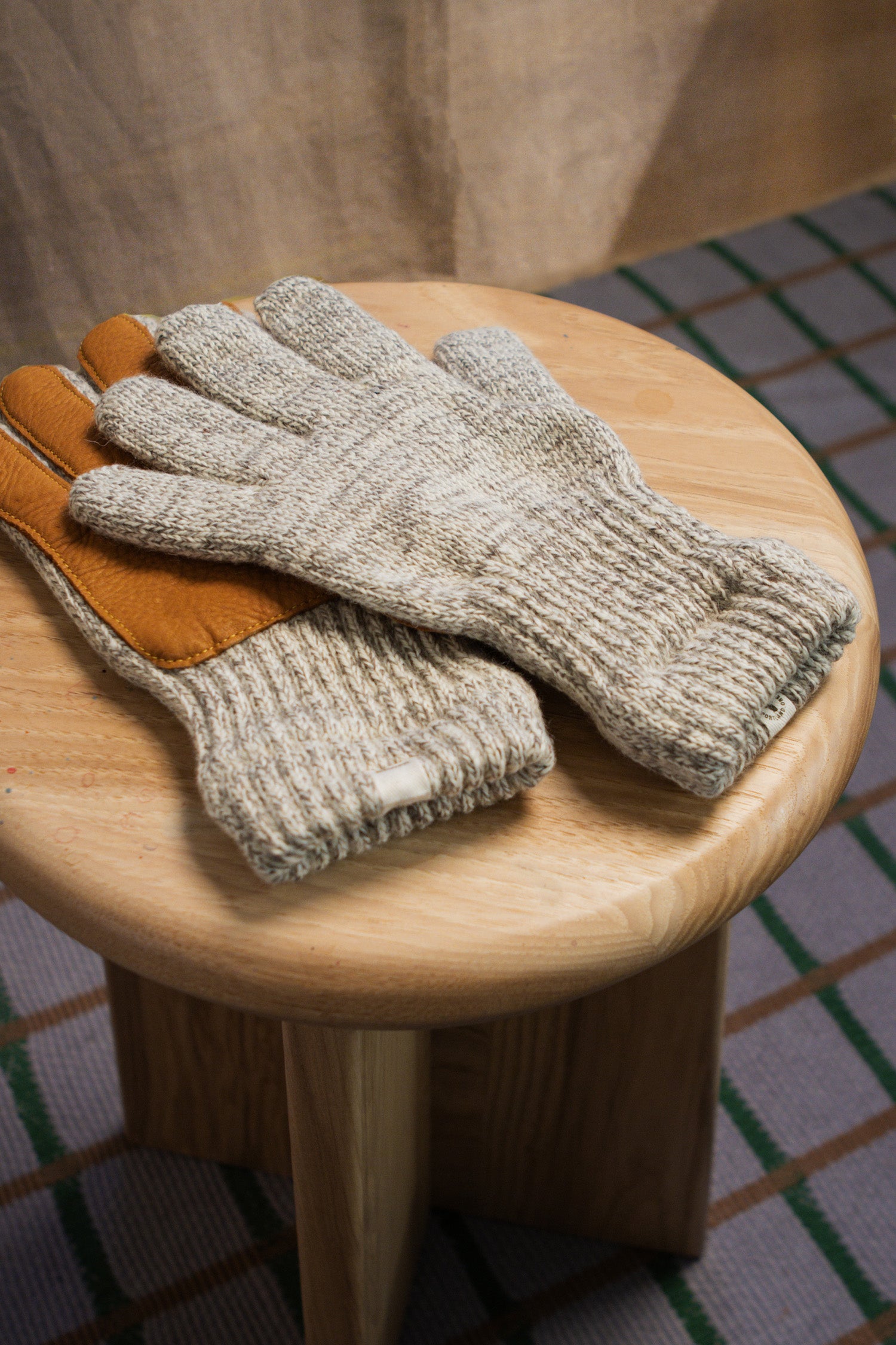 The Bridge & Burn Ragg Wool Lined Gloves in Oatmeal, one with a leather palm, sit atop a round wooden stool with a checked backdrop and beige curtain.