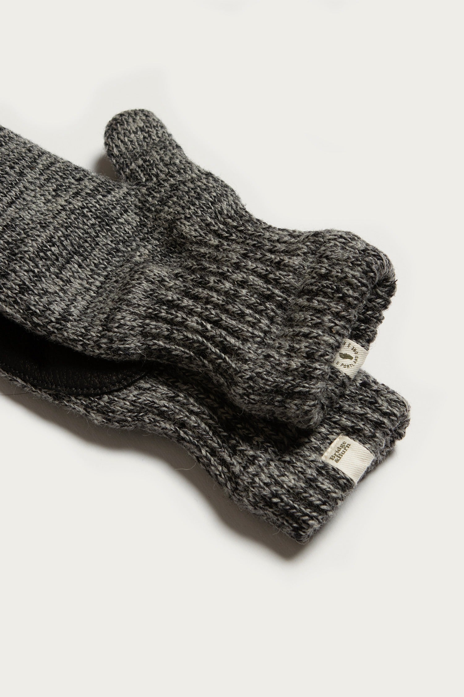 The Bridge & Burn Ragg Wool Lined Mitten in Charcoal features dark grey wool knit, ribbed cuffs, and a small beige fabric tag near the cuff, shown lying on a white surface.