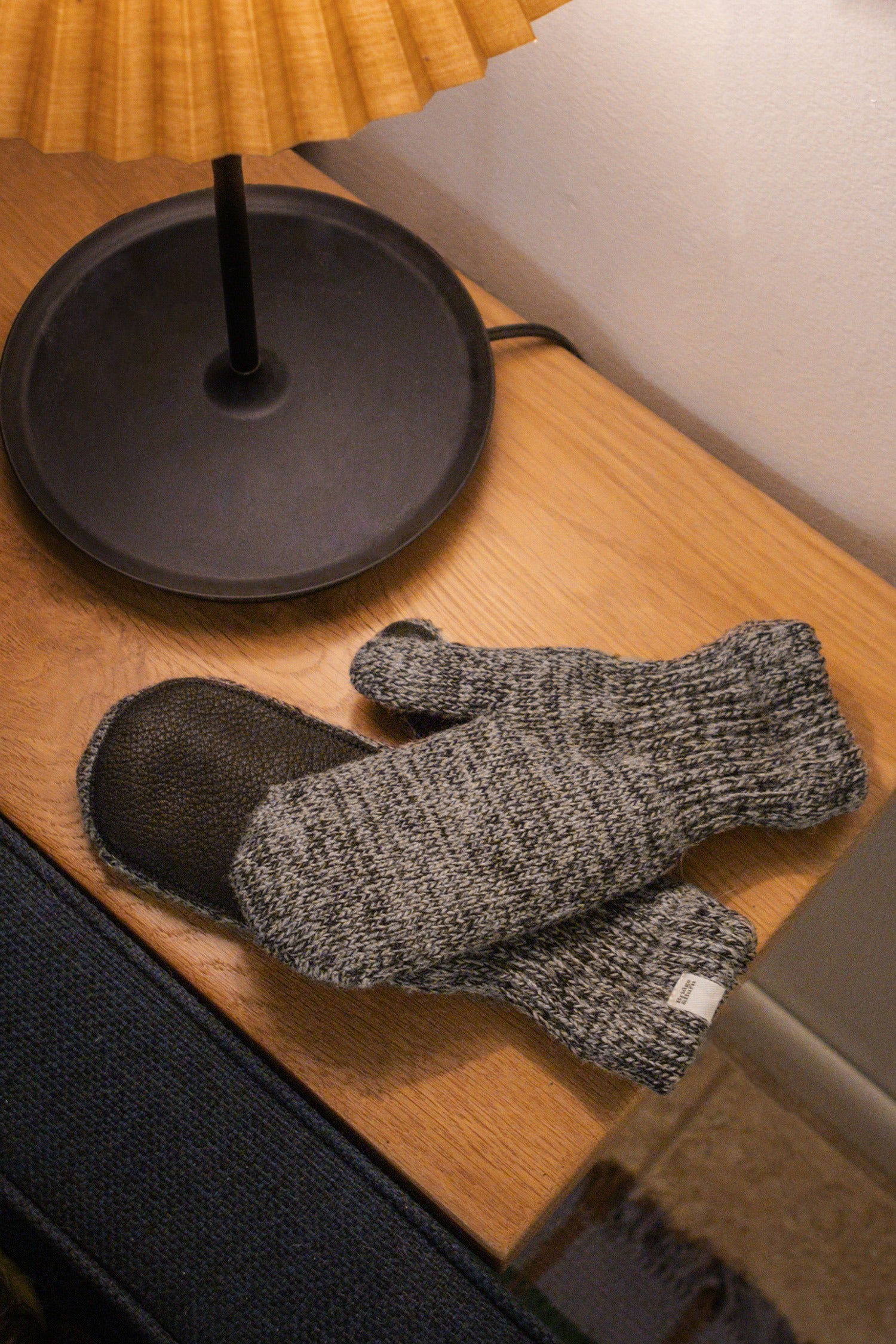 A pair of Bridge & Burn Ragg Wool Lined Mittens in Charcoal sits on a wooden table beside a black lamp with a pleated yellow shade, with part of a blue couch and rug visible in the corner.